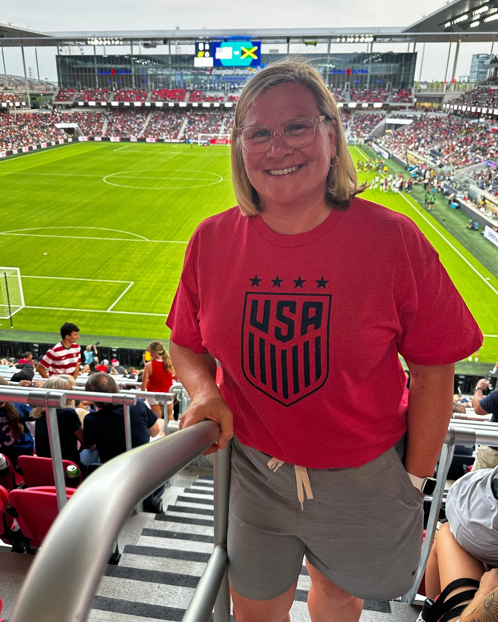 Jen Brooks smiling in a red USA soccer T-shirt in the stands at a soccer stadium