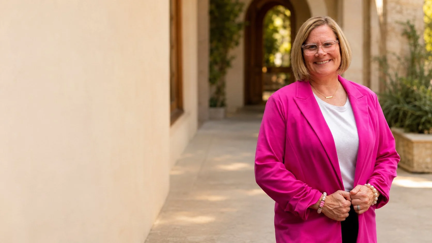 Jen Brooks in a bright pink blazer, standing outdoors by an arched wooden doorway.