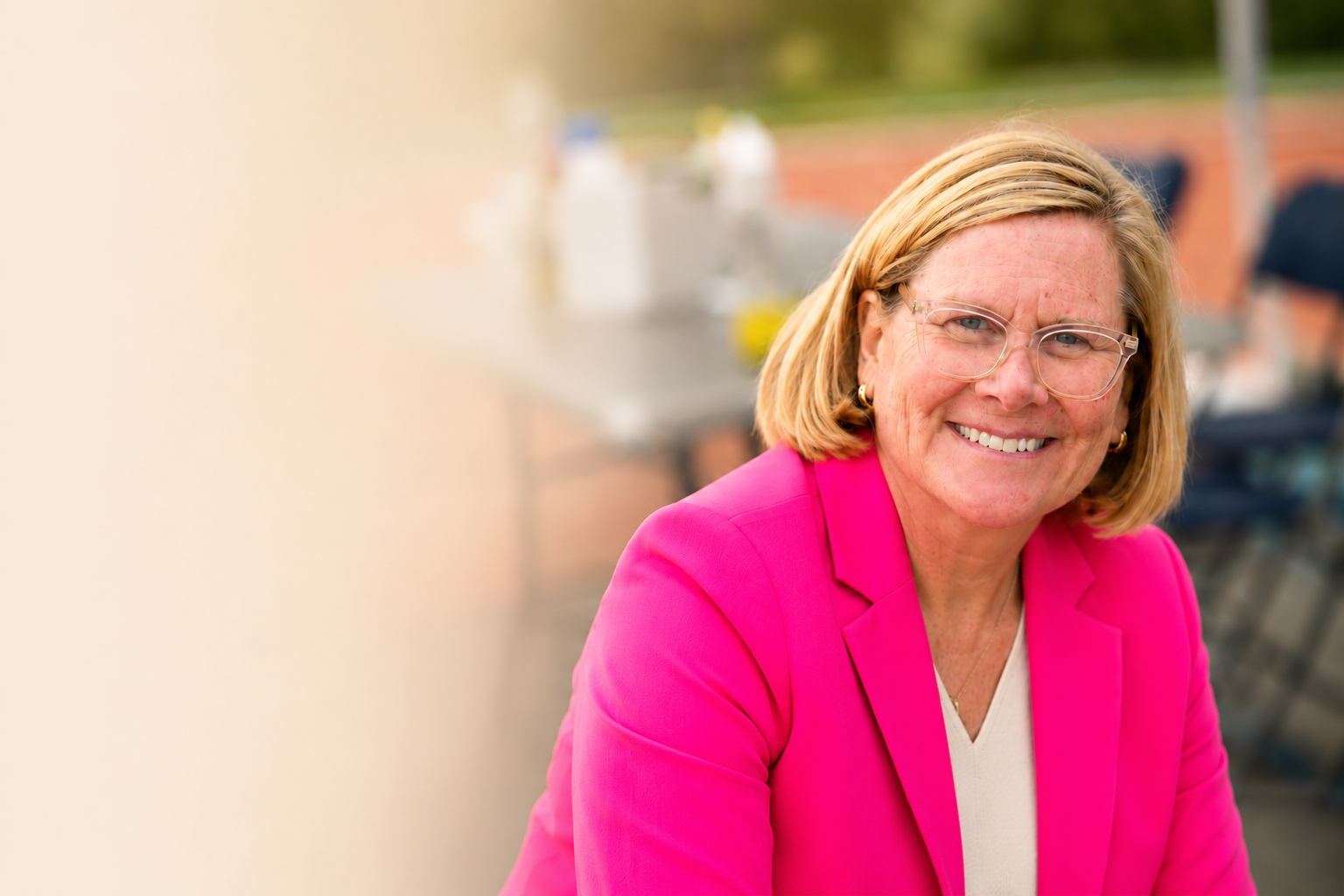 A woman wearing a bright pink blazer and glasses, smiling outdoors with a blurred background of a park or outdoor seating area.