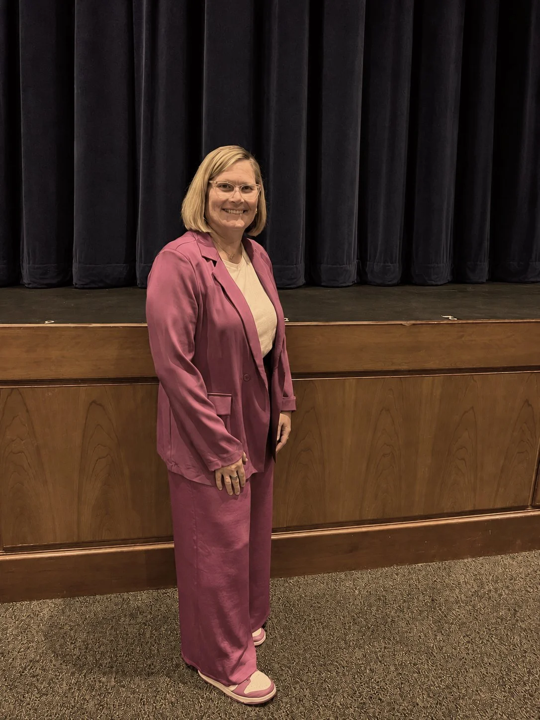Jen Brooks in a pink pantsuit, standing in front of a stage before speaking.