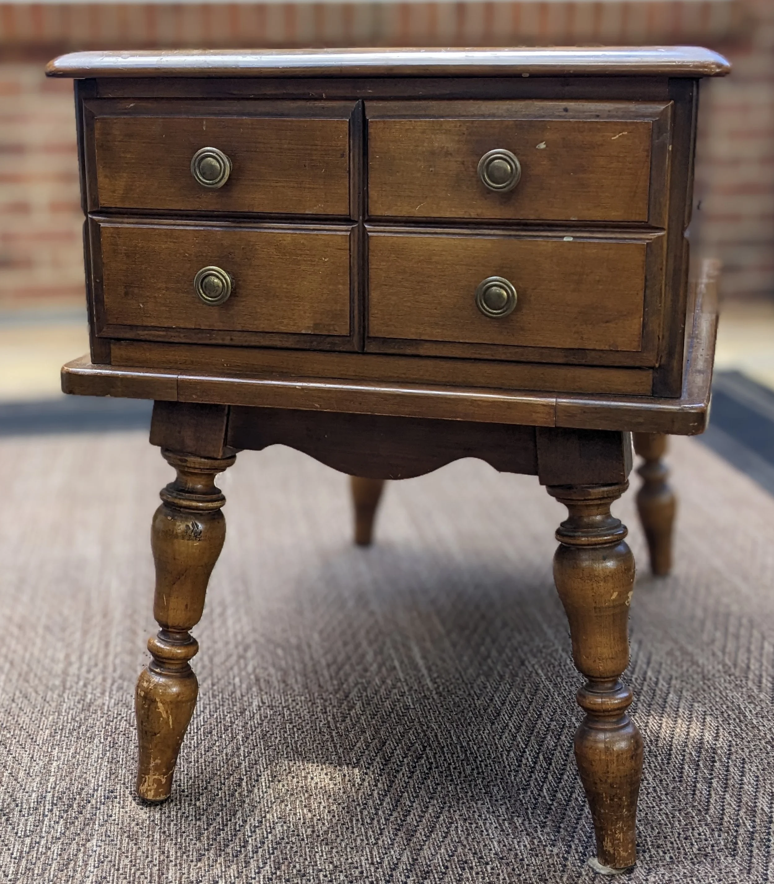 A small vintage wooden table with four drawers, each with a brass knob, set on a textured grey carpet with a brick wall in the background.