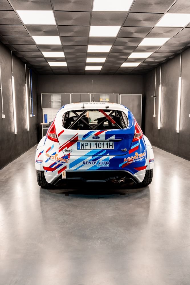 Carro de competição com decoração em azul, vermelho e branco dentro de uma garagem com paredes e teto pretos, com matrícula polaca e vários autocolantes de patrocinadores, incluindo 'Augustinho' e 'Reno Auto'.
