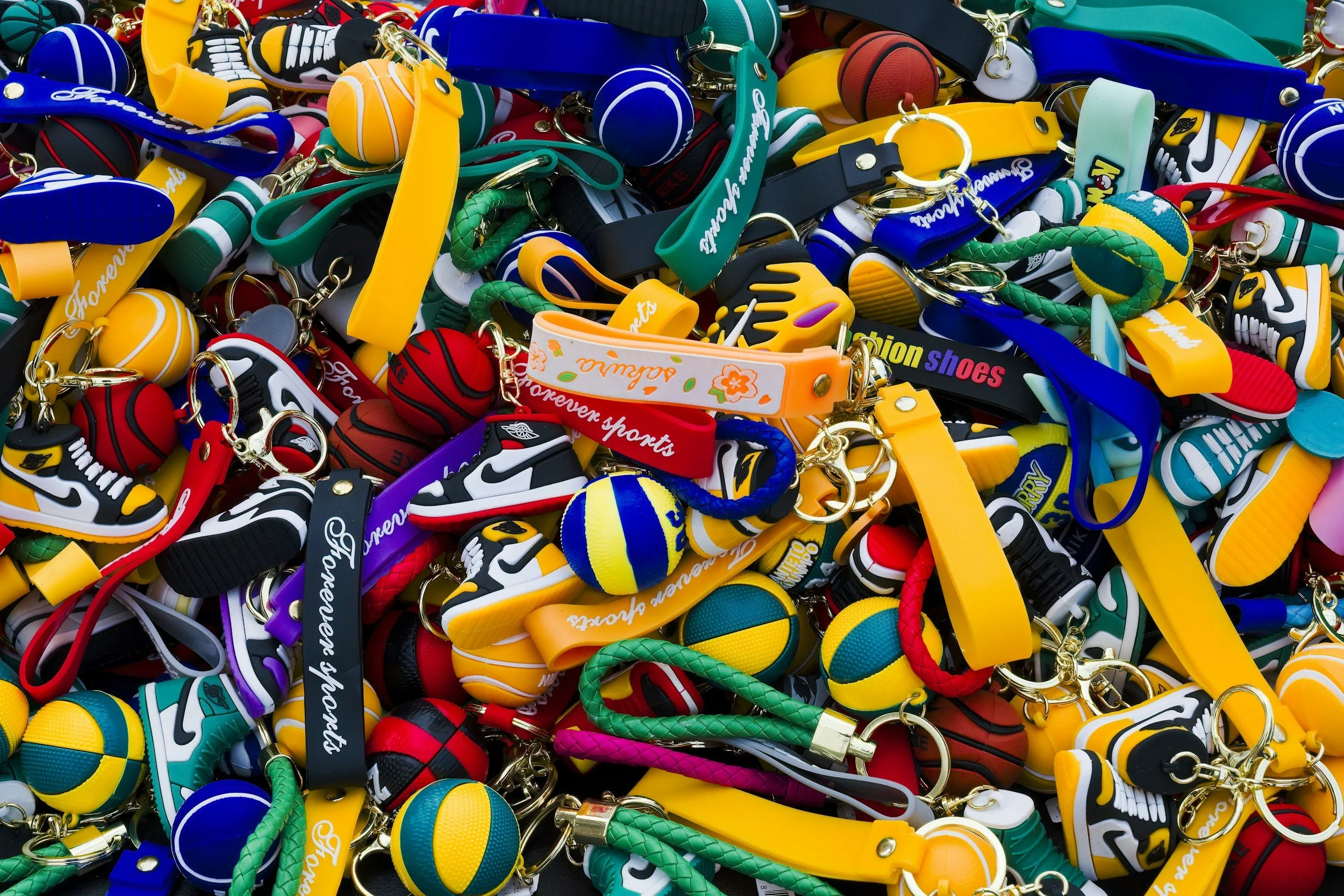 Grande conjunto de porta-chaves coloridos com temática desportiva, incluindo bolas de basquetebol e sapatilhas, em tons de amarelo, vermelho, verde, azul e preto.