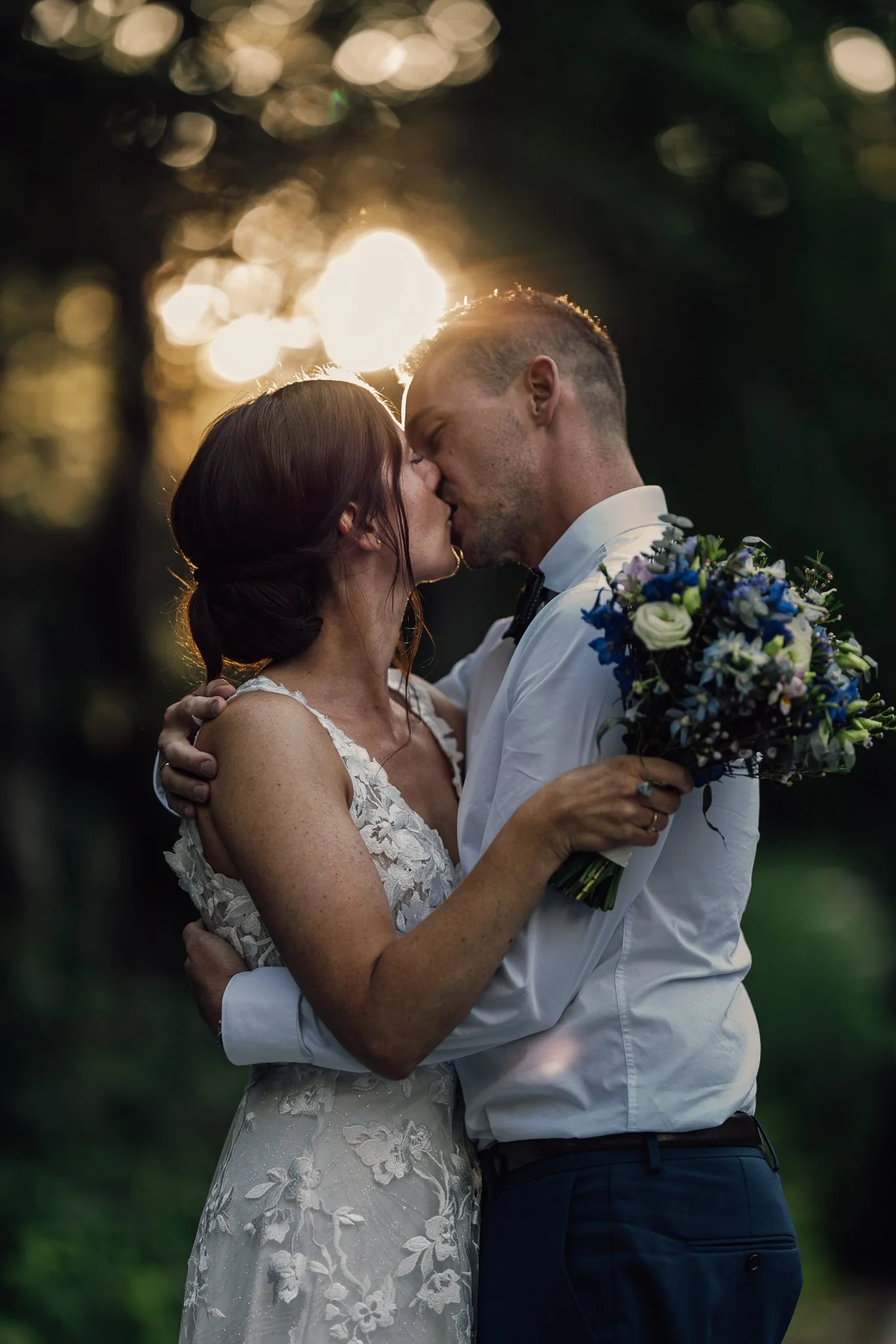 Et brudepar kysser hinanden under solnedgangen i en skov. Kvinden har brunt hår i en opadvendt frisure, og mannen har kort, lyst hår. De holder hinanden i armene, og kvinden holder en blomsterbuket.