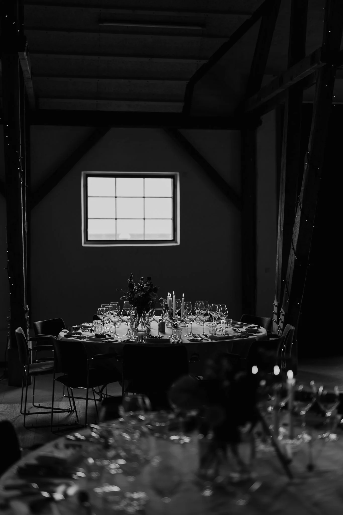 Et rundt bord dækket med vin- og drikkeglas, lys og blomster, i et mørkt rum med en stor vindue og træbjælker, klar til en fest. 