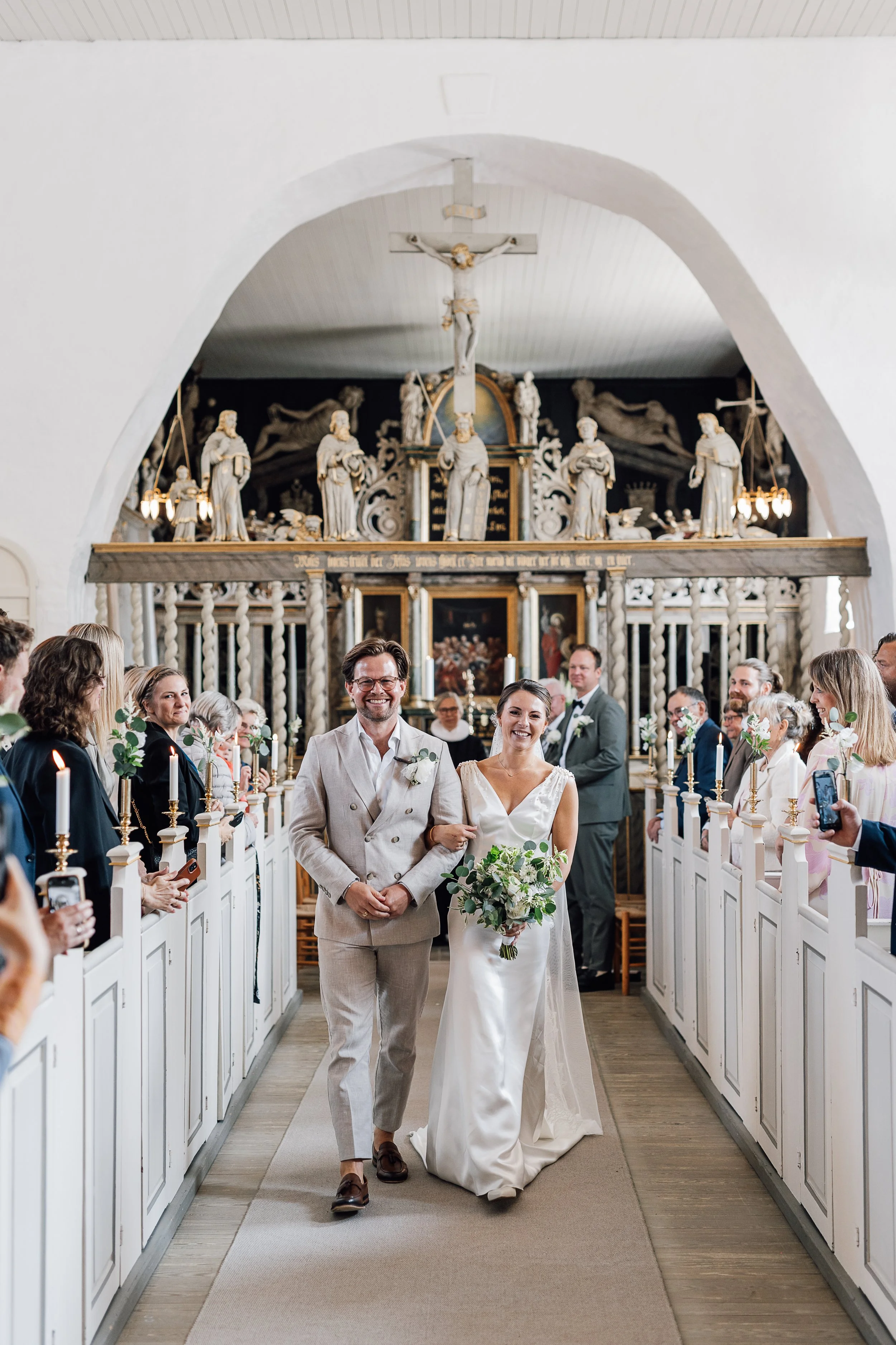 En brudepar går ned ad kirkegulvet under deres bryllupsceremoni, omgivet af gæster, i en kirke med hvide vægge og religiøse figurer på alteret.