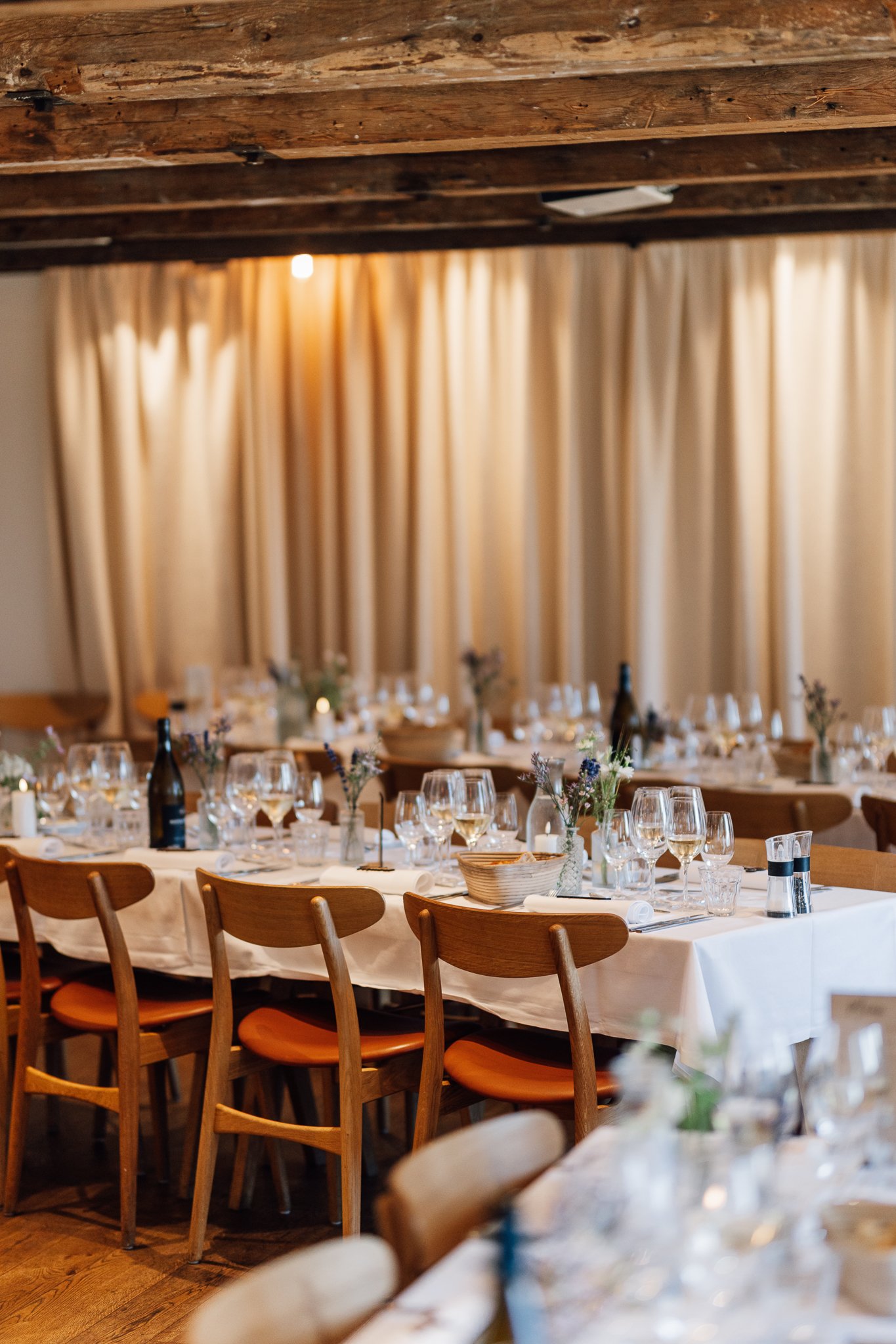 Festmiddag med langbord, vin, blomster og stearinlys i en hyggelig restaurant med beige gardiner og træloft.