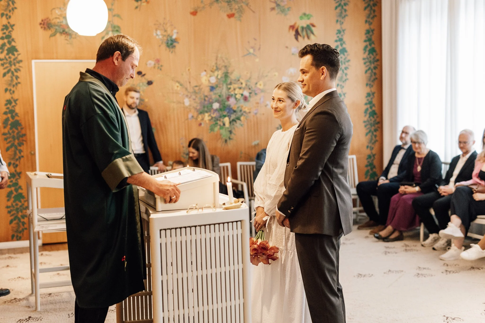 En brud og en brudgom står foran en præst, der holder et børs, i en ceremoni, mens gæster sidder og ser til. Rummet har tapet med blomstermønster.