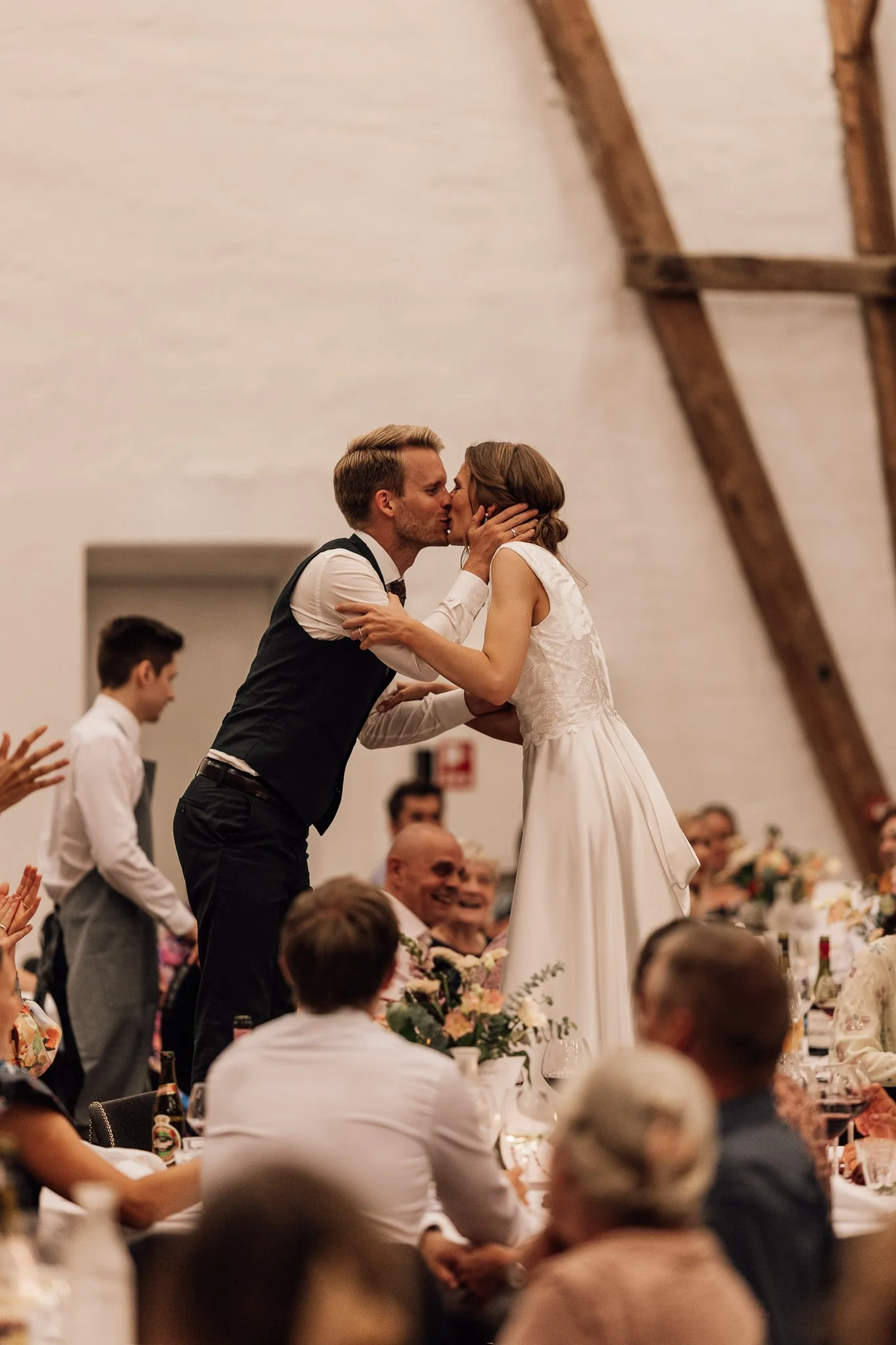 Et par kysser hinanden under en bryllupsfest, omgivet af glade gæster siddende ved langborde med blomster og drikkevarer.