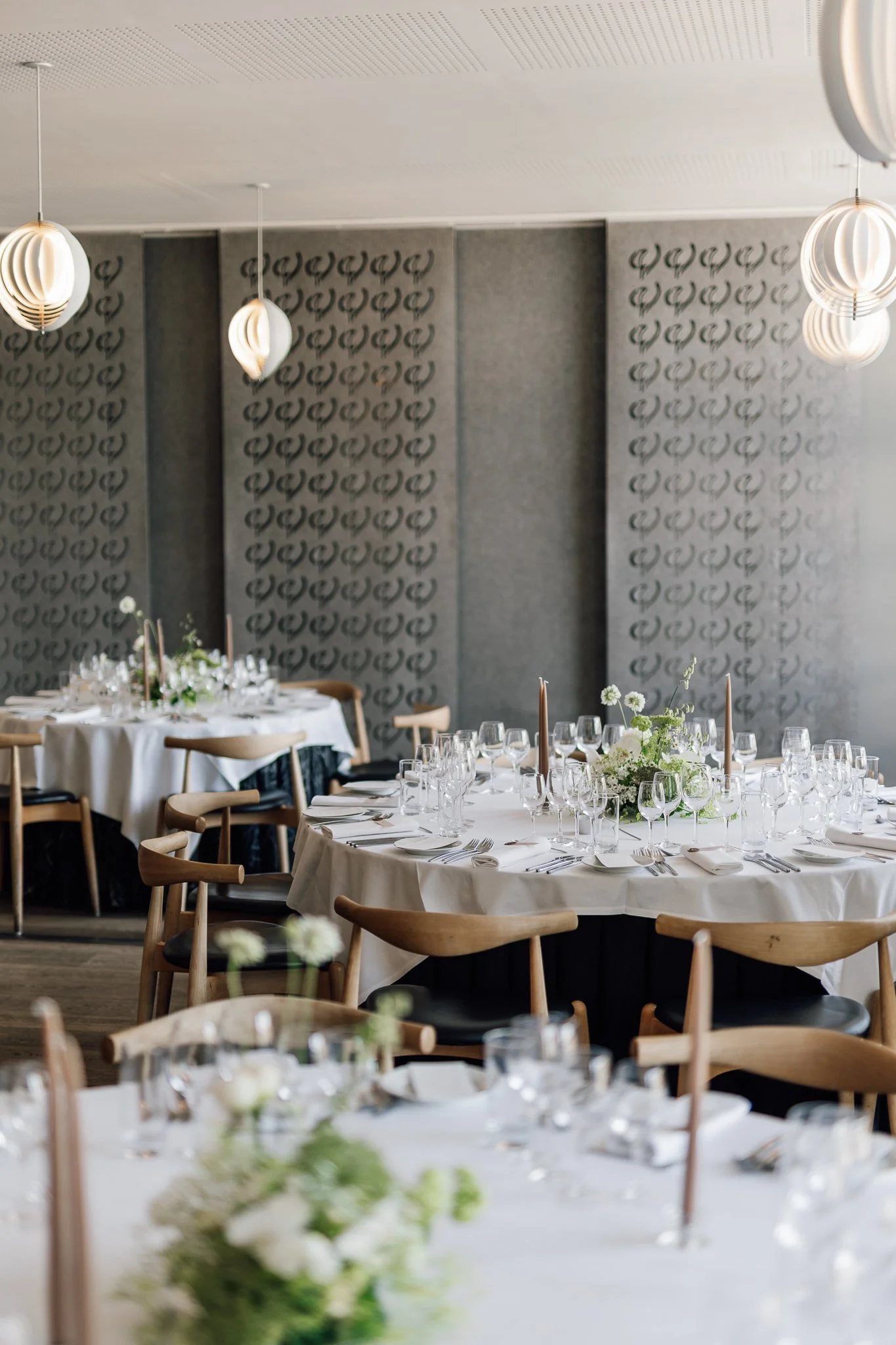 Elegant festlokale med rundborde dækket med hvide duge, blomsterdekorationer og stearinlys, samt moderne pendellamper i loftet.
