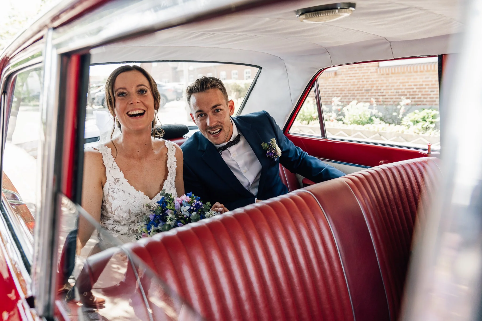 Et brudepar sidder i en rød vintagebil, hvor kvinden bærer en hvid brudekjole og holder en blomsterbuket, mens manden er klædt i en blå jakkesæt med butterfly.