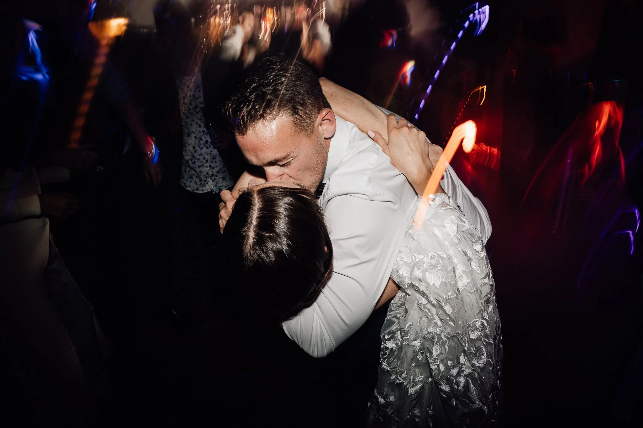 To personer i formelt tøj danser tæt sammen, omgivet af personer i festligt tøj med farverige lysstriber i baggrunden.