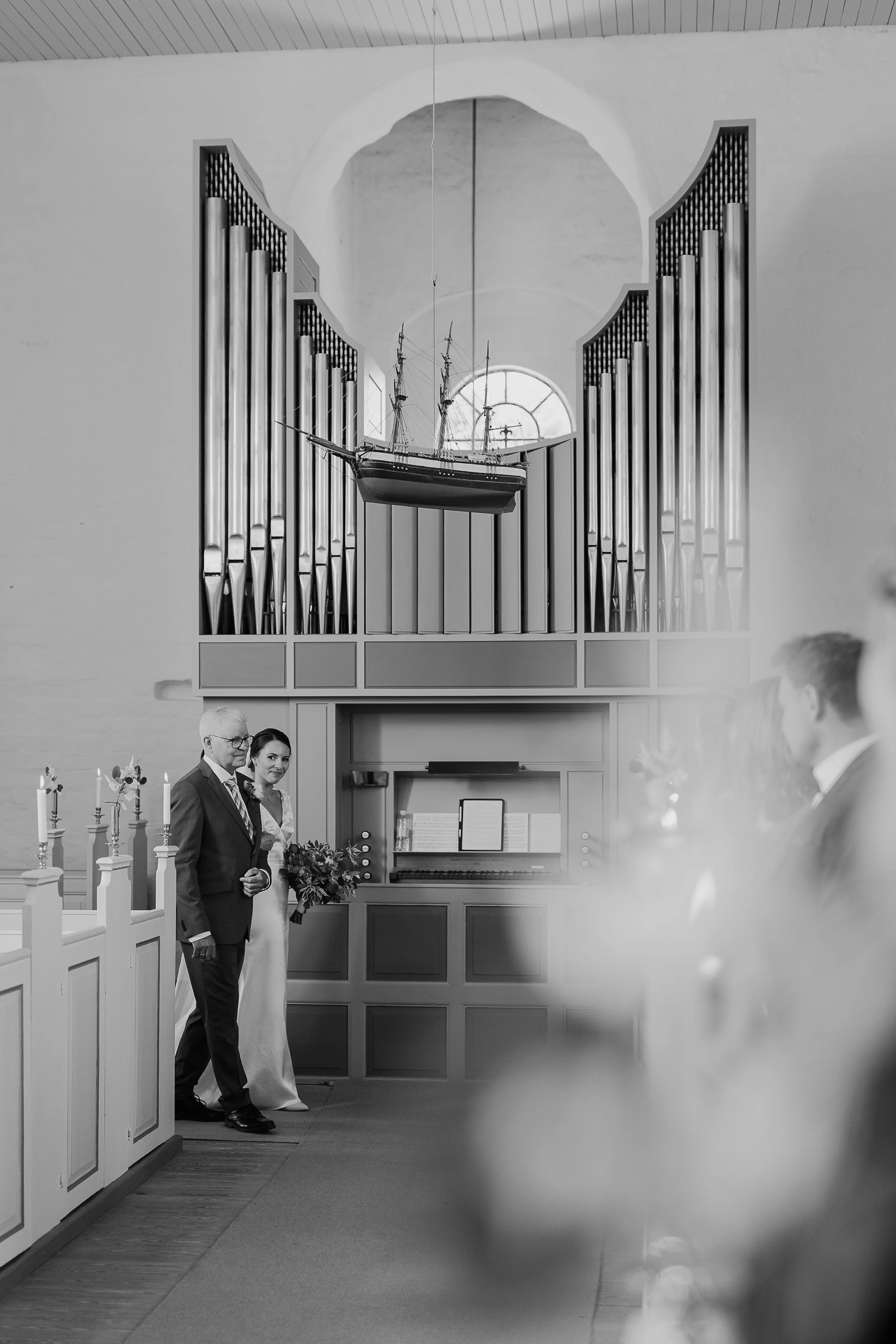 Sort-hvid fotografi af et bryllupssted med en couple foran kirkens alter, der står ved en hvid glaseret kor med en modelskib hængende fra loftet.