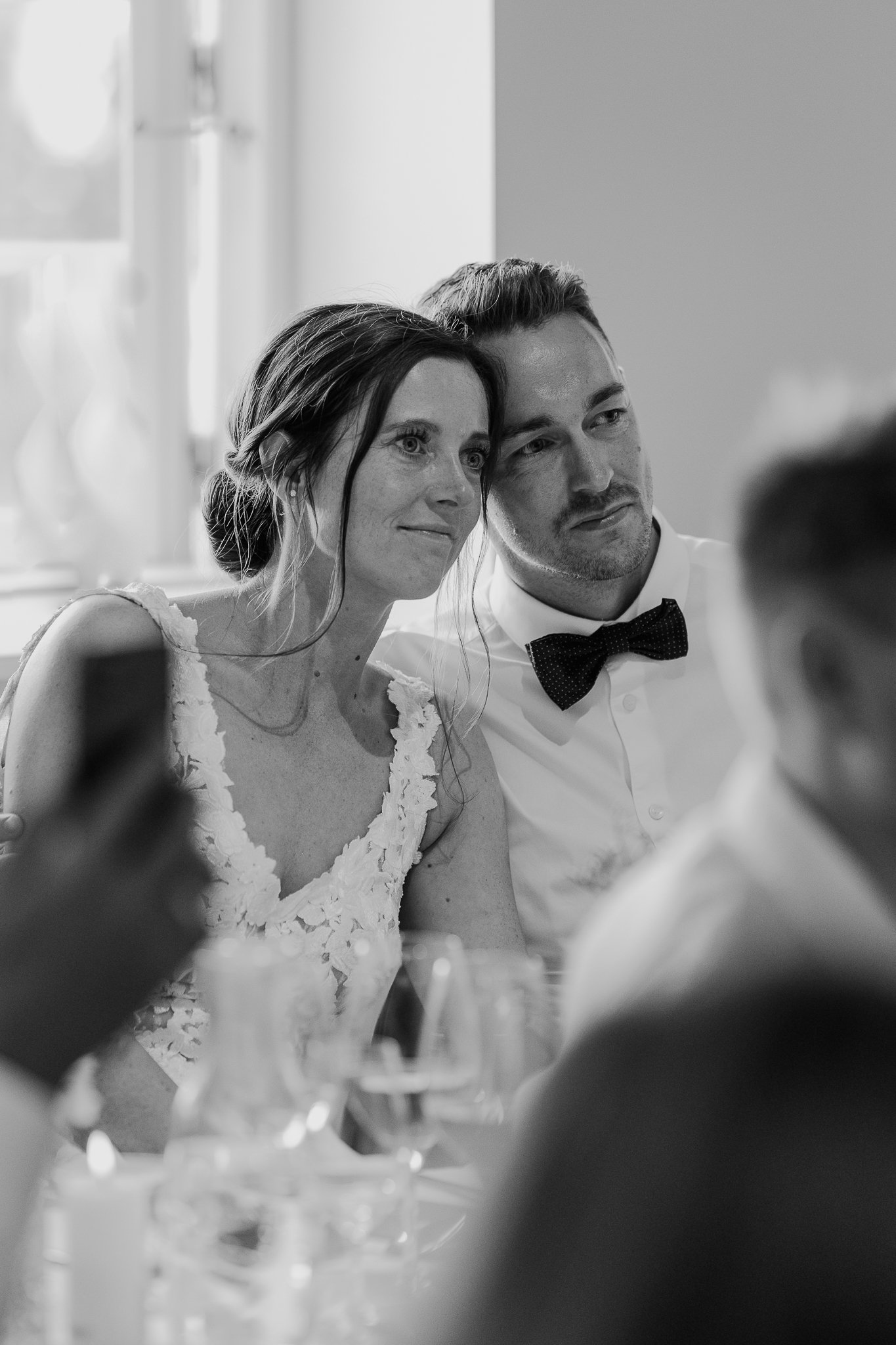 Sort-hvid foto af et par ved et bord, sandsynligvis til en formel begivenhed, med slidte ansigter, udtrykker nærhed og hygge, med glas og blomster på bordet.