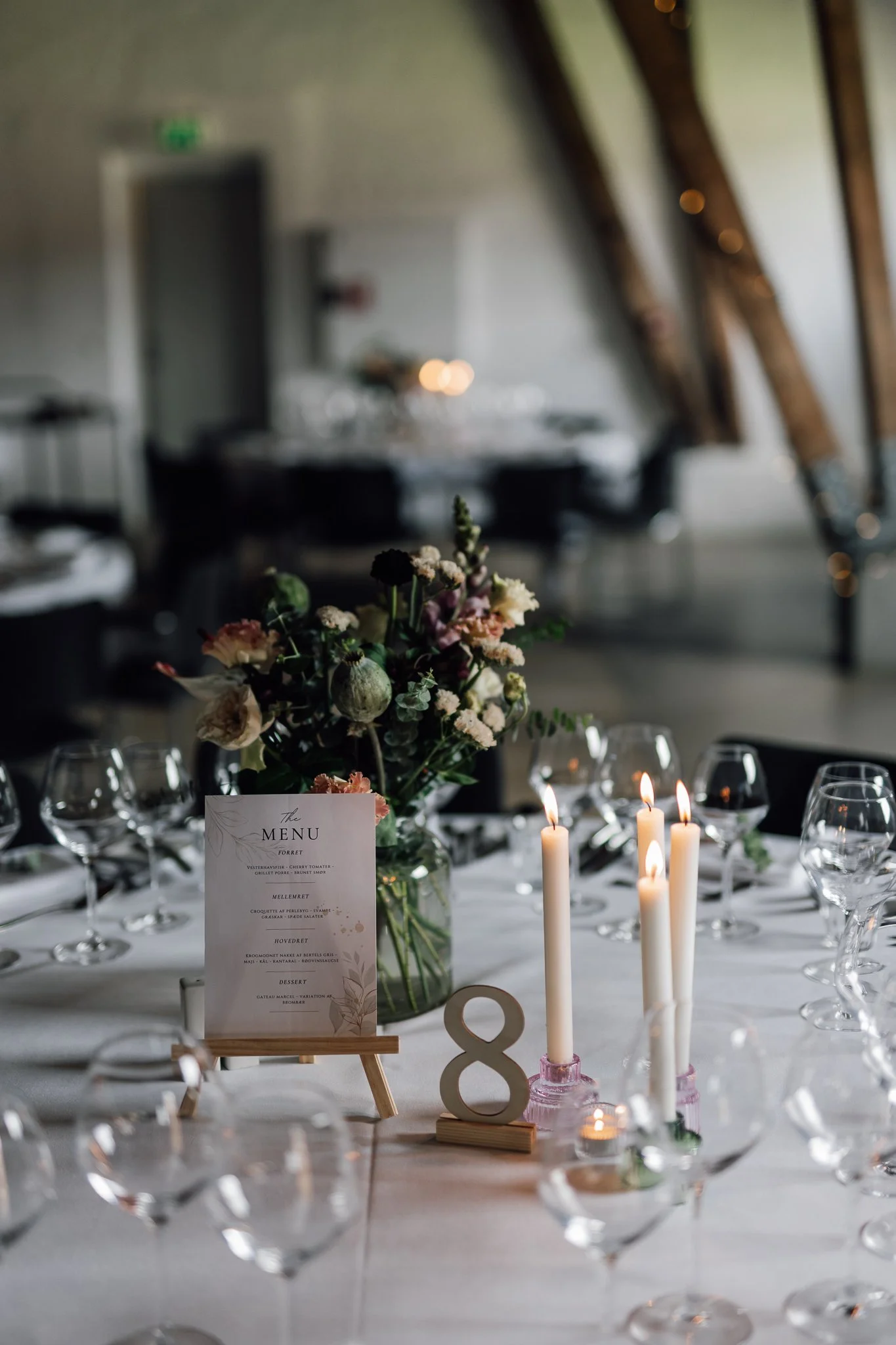 Borddækning med blomster, tændte stearinlys og en menu, klar til en formel middag i et elegant lokale med træbjælker i loftet.