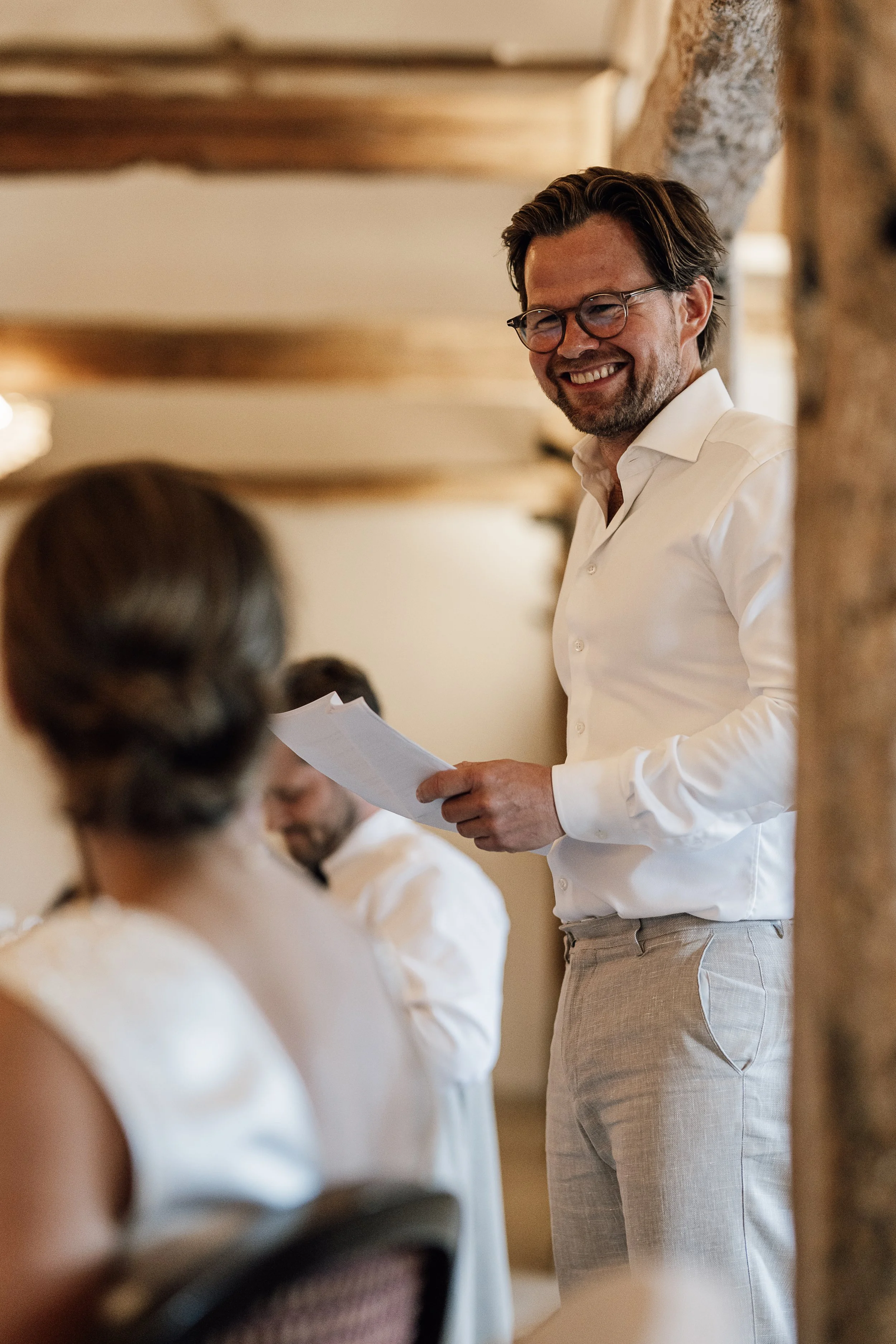 En mand med brunt hår, briller og skæg smiler, mens han holder et stykke papir og står foran flere kvinder, der er klædt i hvide kjoler, i et rum med trævægge.
