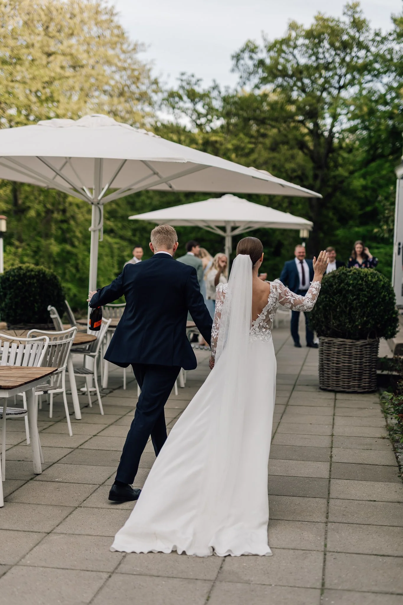En brud og en brudgom danser udendørs på en veranda med parasoller, omgivet af grønne træer og tilskuere i baggrunden.