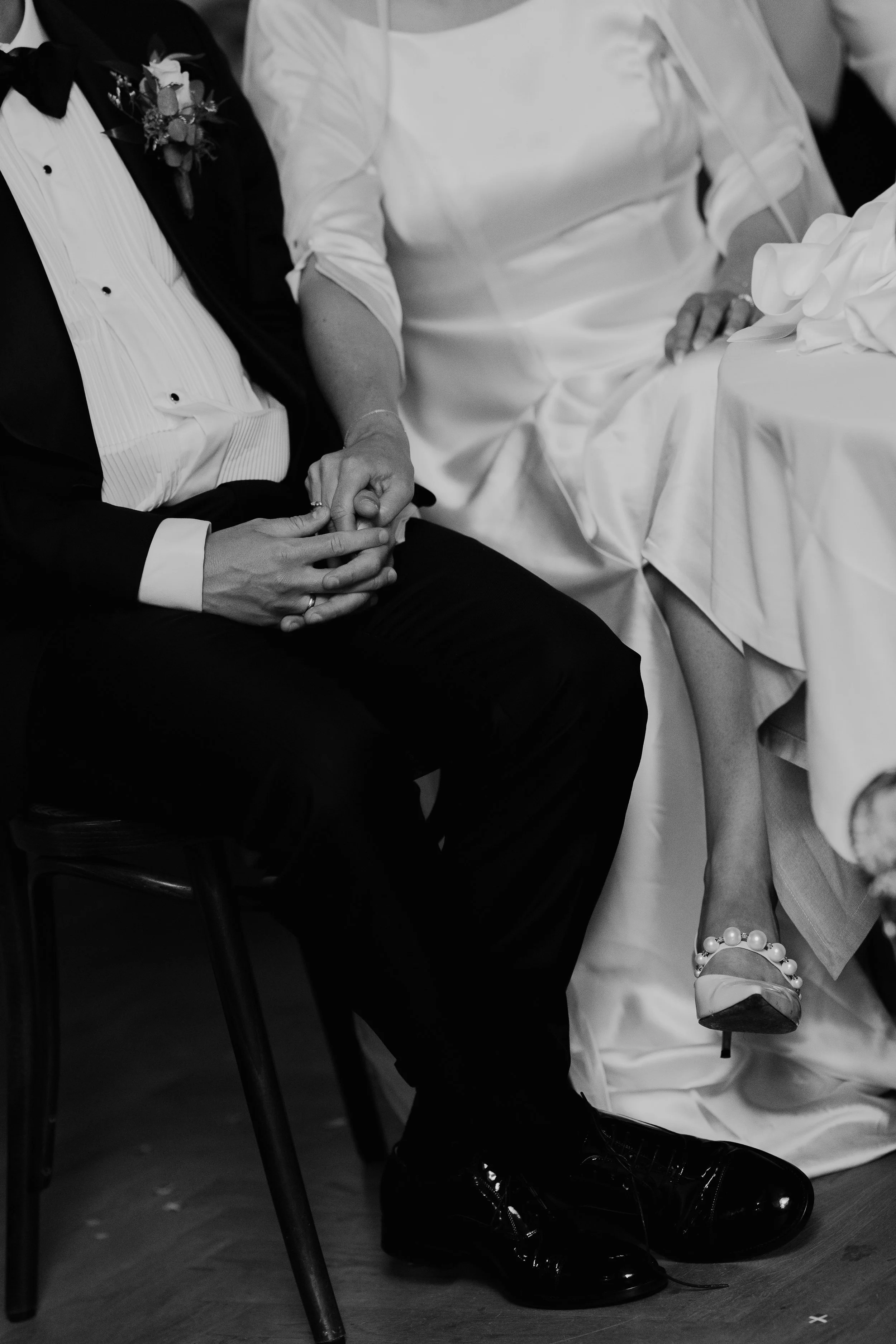 To personer sidder tæt sammen, hånd i hånd, ved en festlig begivenhed. En person har en hvid kjole og armbånd, den anden person har en sort smoking med boutonnière.