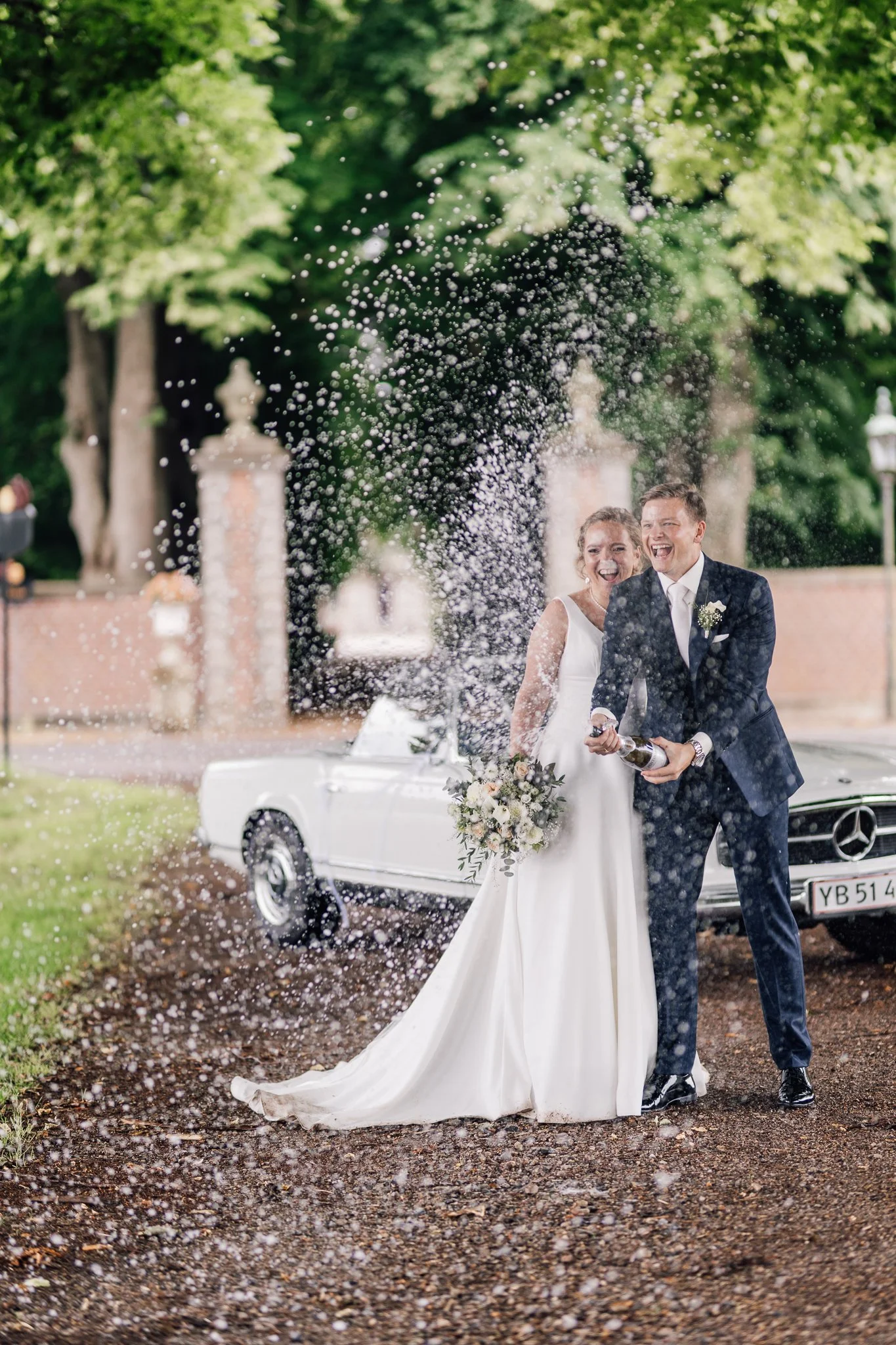 En brud og brudgommen smiler og skåler med champagne foran en hvid bil efter deres bryllup.