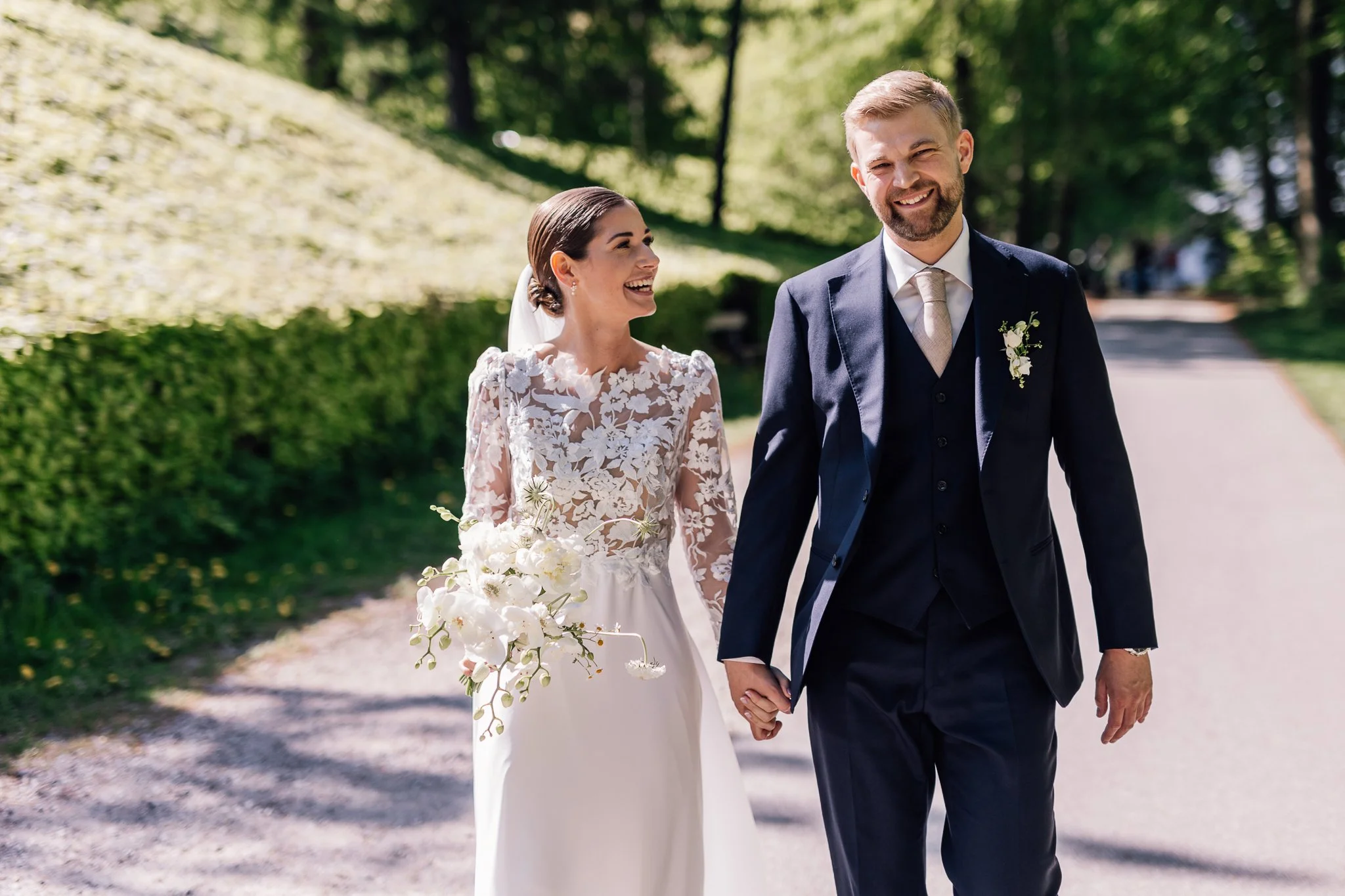 Et nygift par går hånd i hånd i en park, kvinden bærer en brudekjole og holder en blomsterbuket, mens manden er iført en mørk skreddersyet jakkesæt.