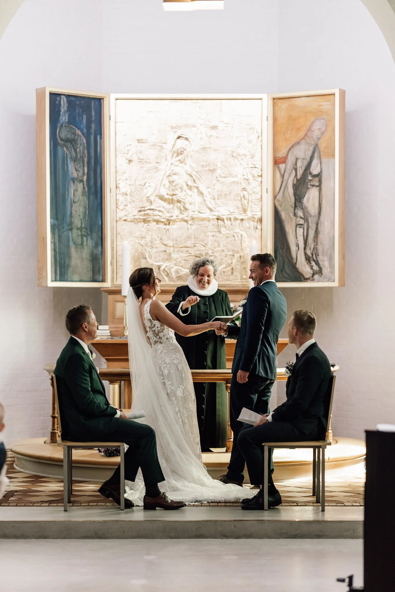 En bryllupsceremoni i en kirke med en brud og en gommen, der holder hænder, omgivet af deres følgere. Præsten står foran dem, og hænger på væggen bagved er tre store kunstværker.