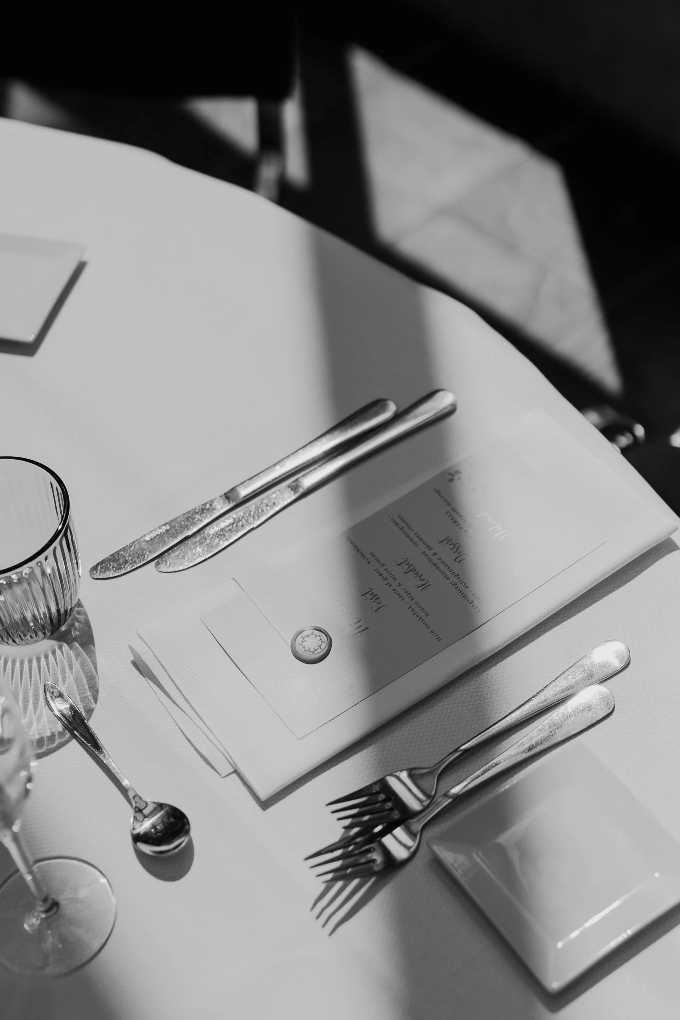 Sort-hvid billede af et bord med sølvbestik, glas, en tallerken med en kuvertlappetelig festmenu, og en serviet, sat op til bryllup.