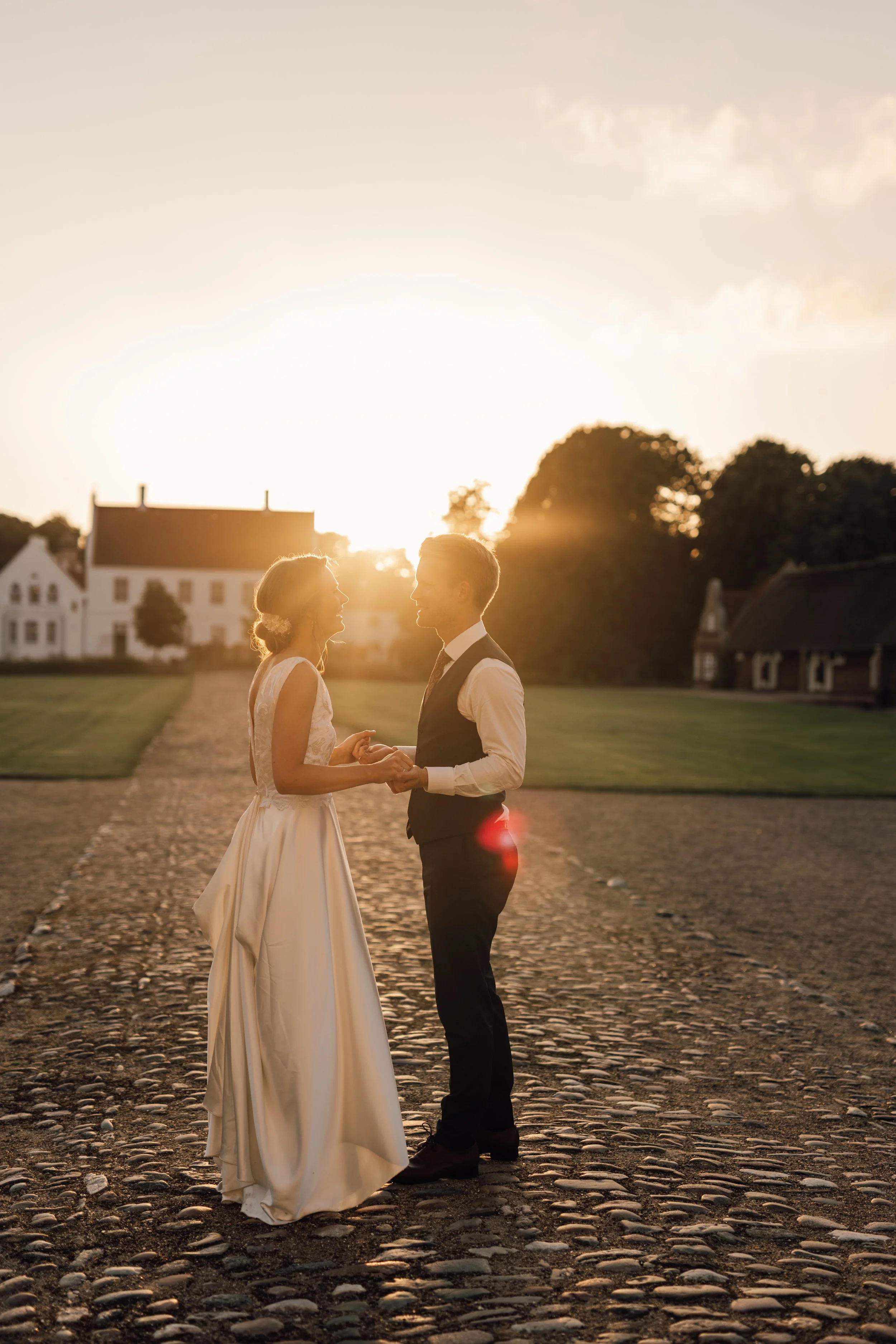 En brud og en brudgom står på en grusbelagt vej ved solnedgang, holder hinanden i hænderne og ser hinanden i øjnene, omgivet af grønne træer og gamle huse.