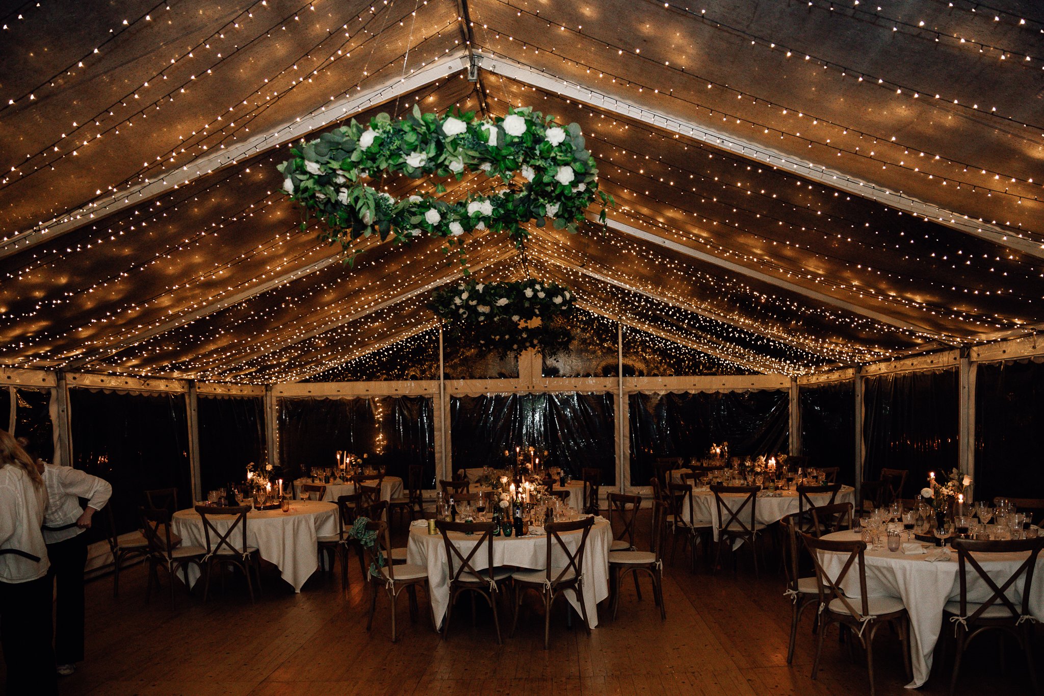 Indendørs festlokale med rundborde dækket med hvide tableauer, stearinlys og blomsterdekorationer, og loftet er pyntet med spredte små lysstrimler og en stor blomsterkrans.
