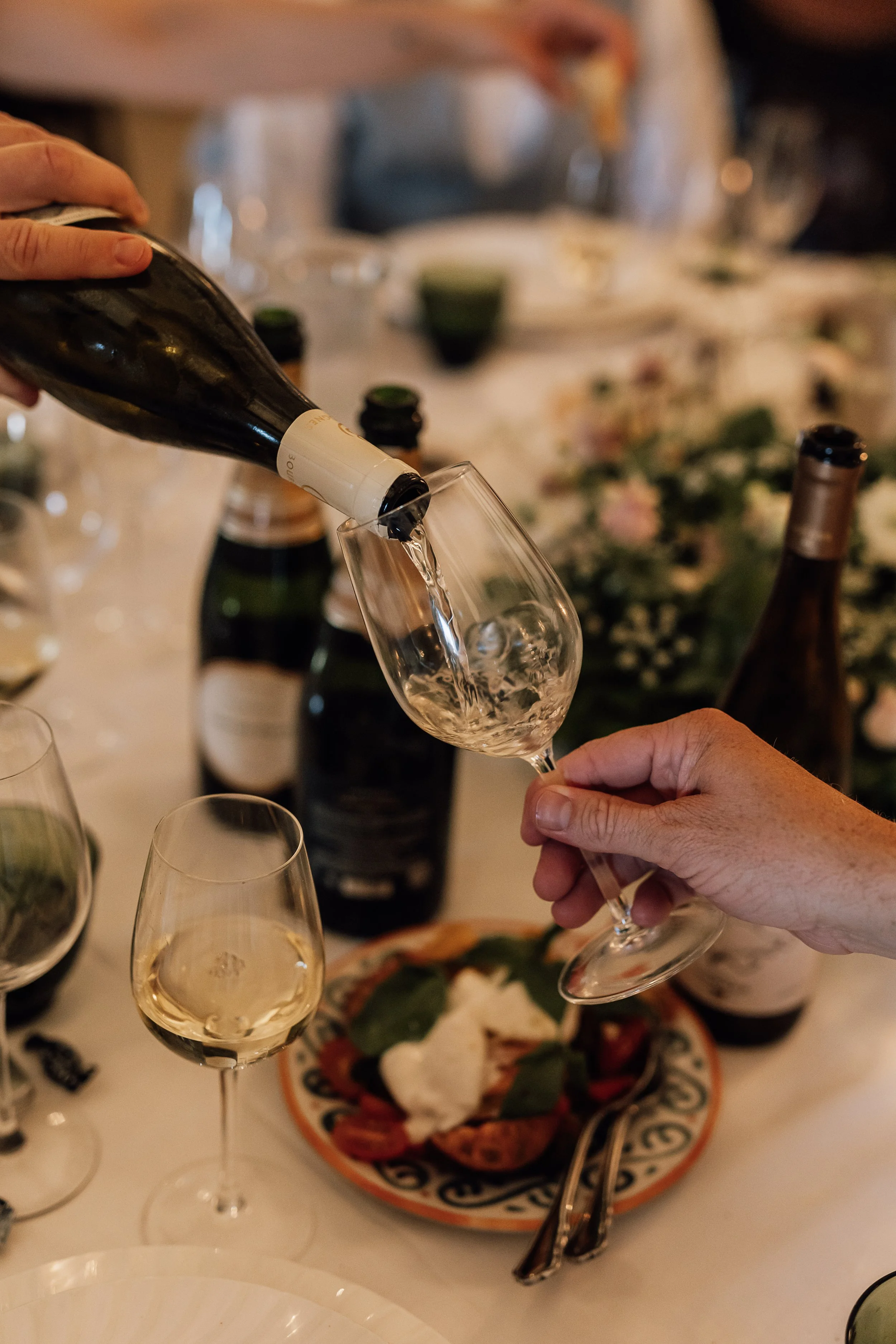 En person hælder mousserende vin i et vinglas ved et bord dækket med flere tomme og fyldte vinglas, ølflasker og en tallerken med mad.