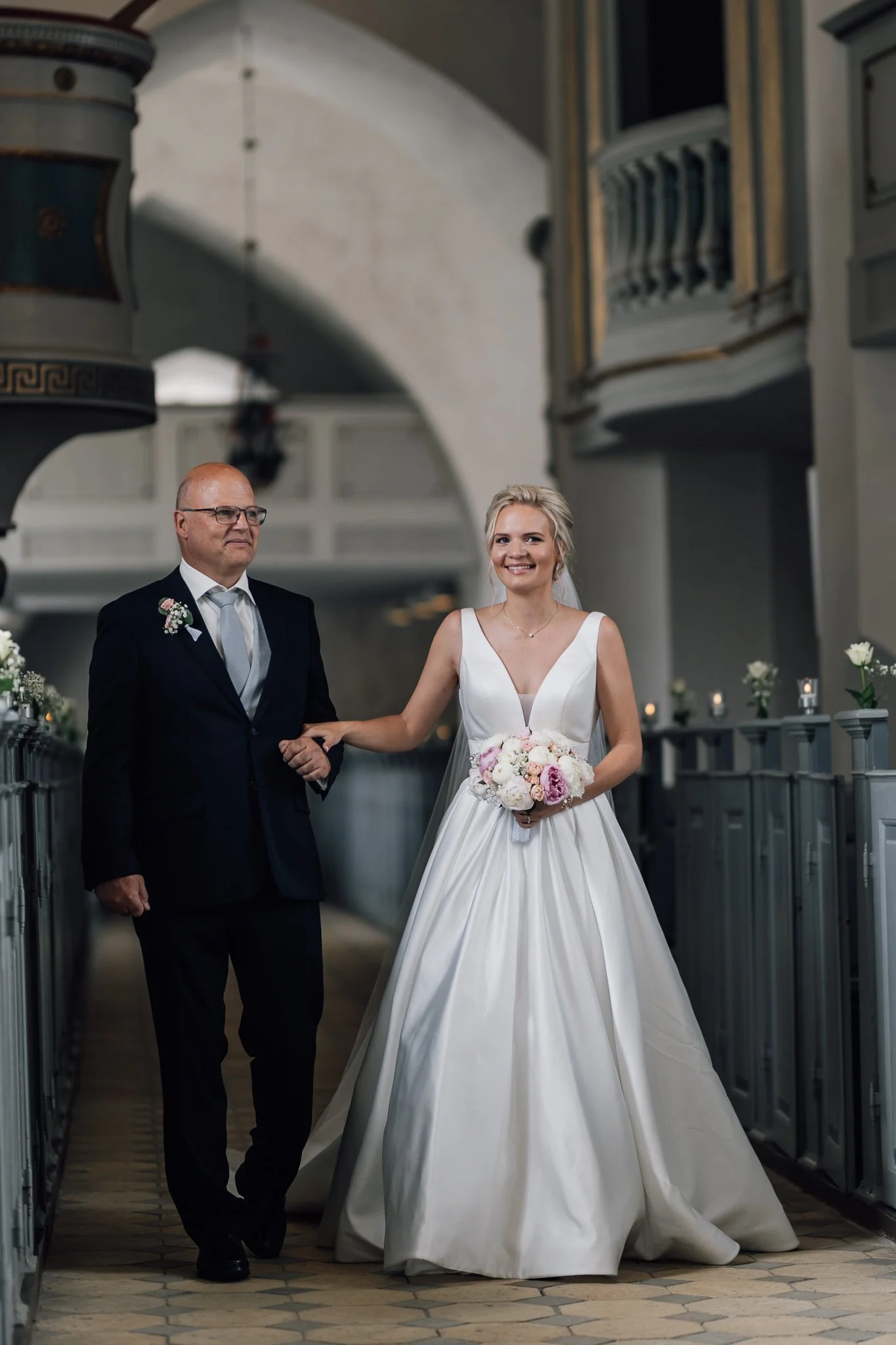 En mand og en kvinde på vej ind i kirke, manden har smoking og kvinden i brudekjole med buket i hånden, de smiler.