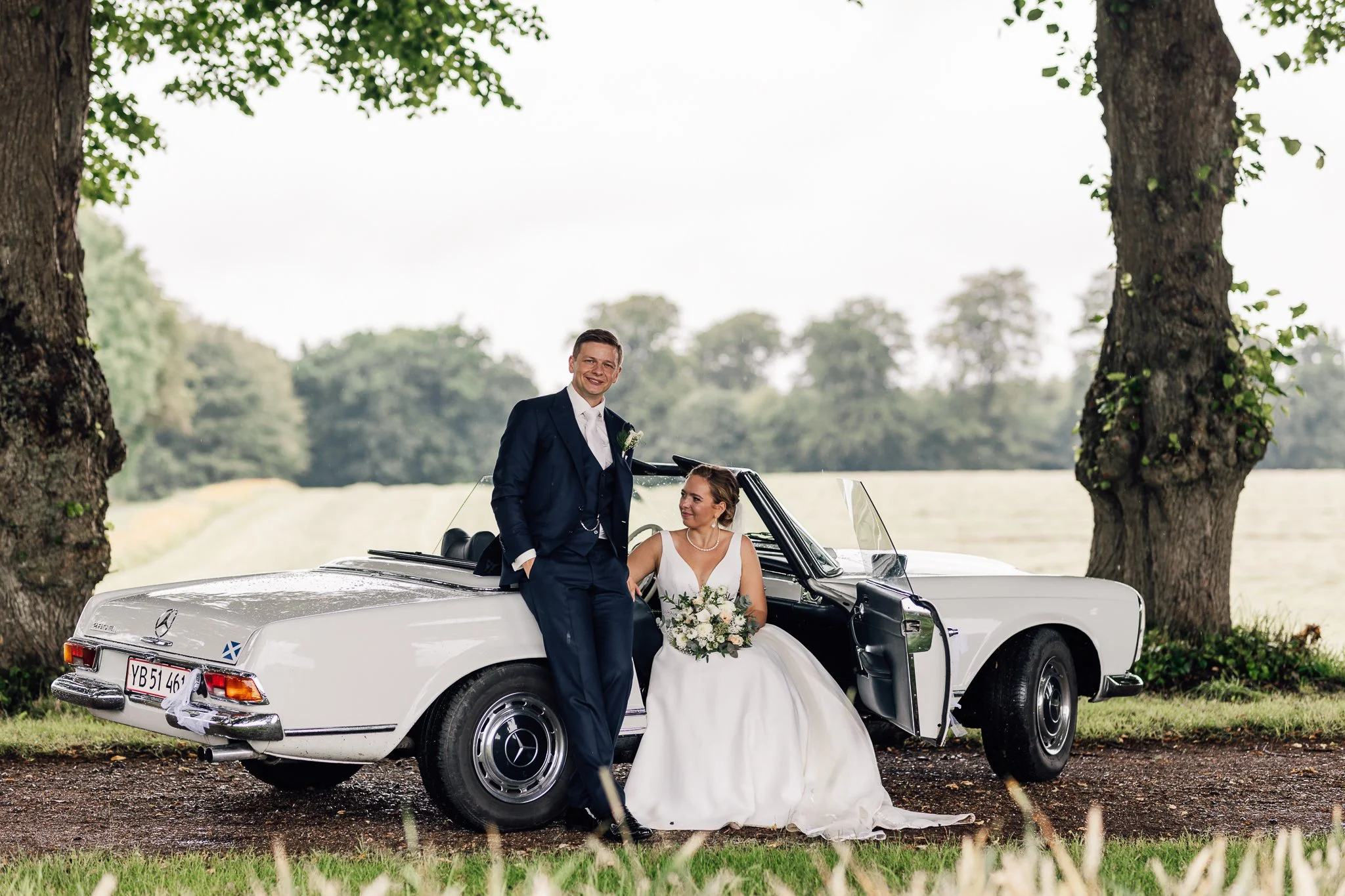 En brud og en brudgom står ved en klassisk hvid Mercedes-Benz cabriolet med en brudebuket, under to store træer i en grønskov. Bruden sidder i bilen, mens brudgommen står med hænderne i lommerne, smilende. Baggrund af åbne marker og træer.