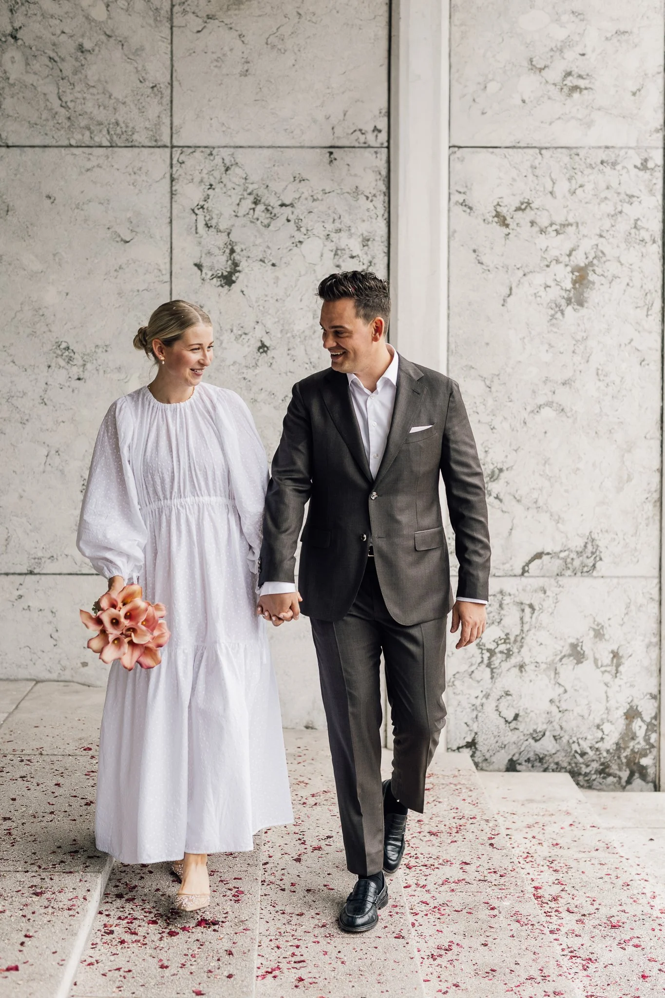 Et lykkeligt brudepar holder hånd ved et bryllup. Kvinden bærer en hvid kjole og holder en buket blomster, mens mænden har en grå habit og sorte sko.