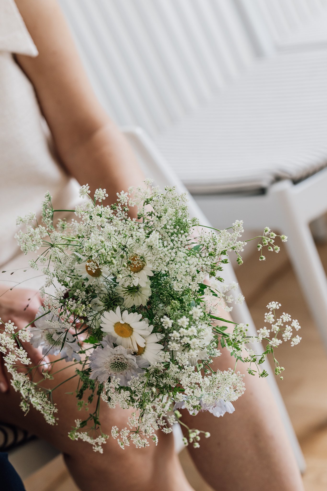 Person, der sidder med benene samlet og holder en blomsterbukett med hvide blomster, herunder mælkebøtter og små hvide blomster, i sofa eller seng i et stue- eller soveværelse.