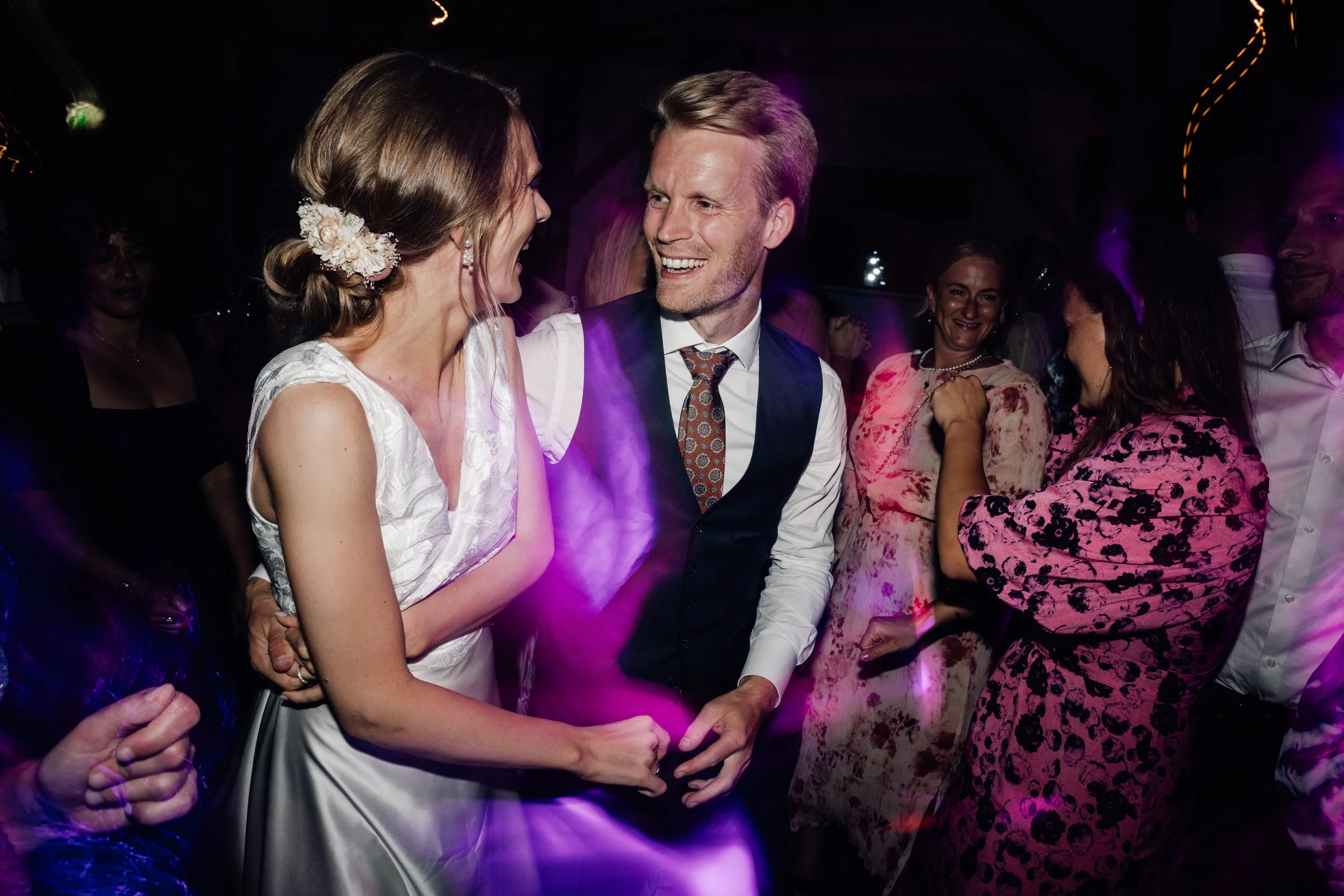 Et bryllupsceremoni med et man & kvinde der smiler og holder hænder, omgivet af familie og venner, i en festlig setting med mørk baggrund og farverig belysning.
