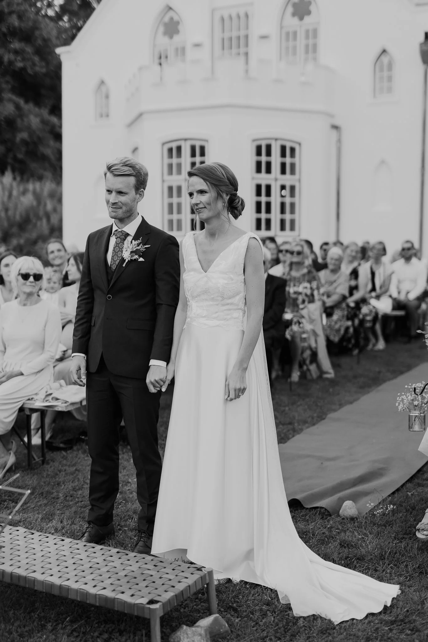 En bryllupsceremoni udenfor foran en hvid bygning, hvor en mand i sort jakkesæt og en kvinde i en lang, hvid kjole står og holder hinanden i hænderne, omgivet af tilskuere.