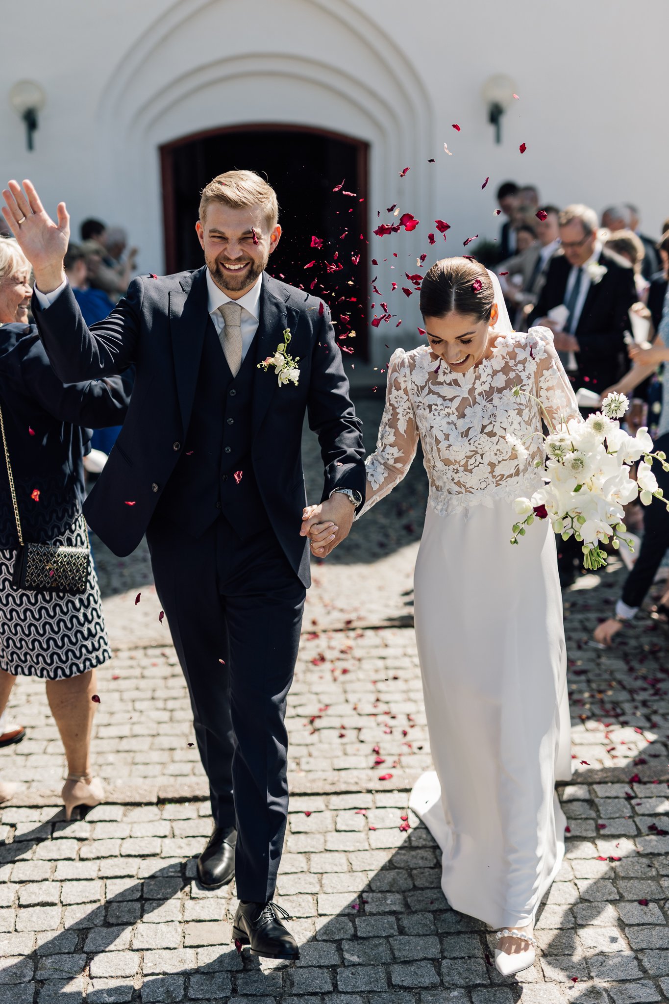 En brud og en gom går hånd i hånd ud af kirken, omgivet af gæster, mens der kastes med blomsterblade, og solen skinner.