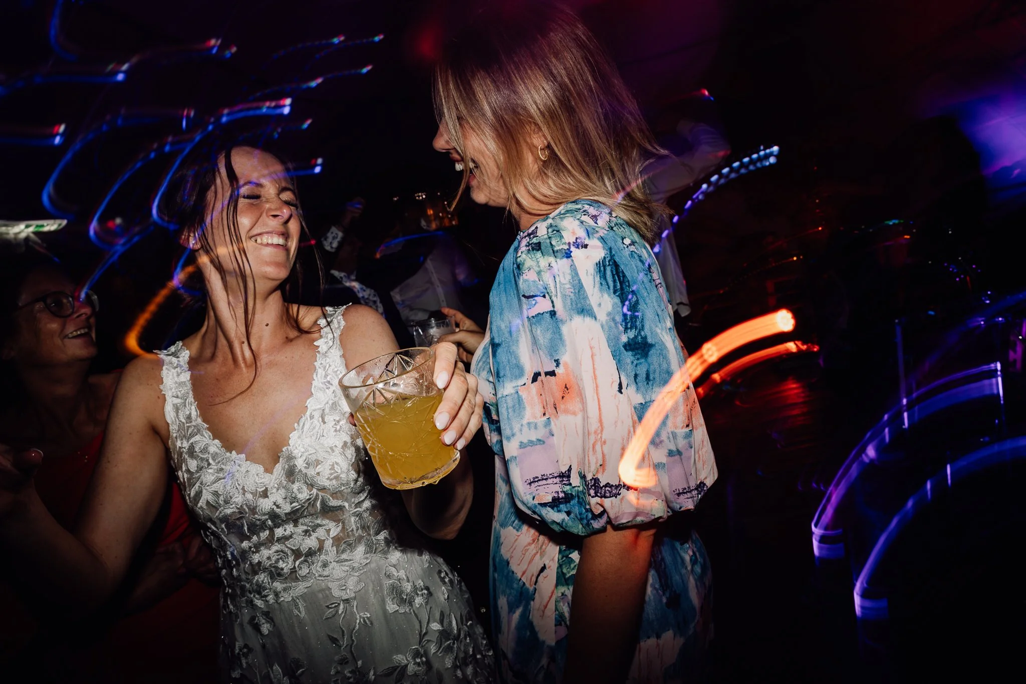 To kvinder danser og har det sjovt på en fest, en holder et glas med en drink, begge smiler.