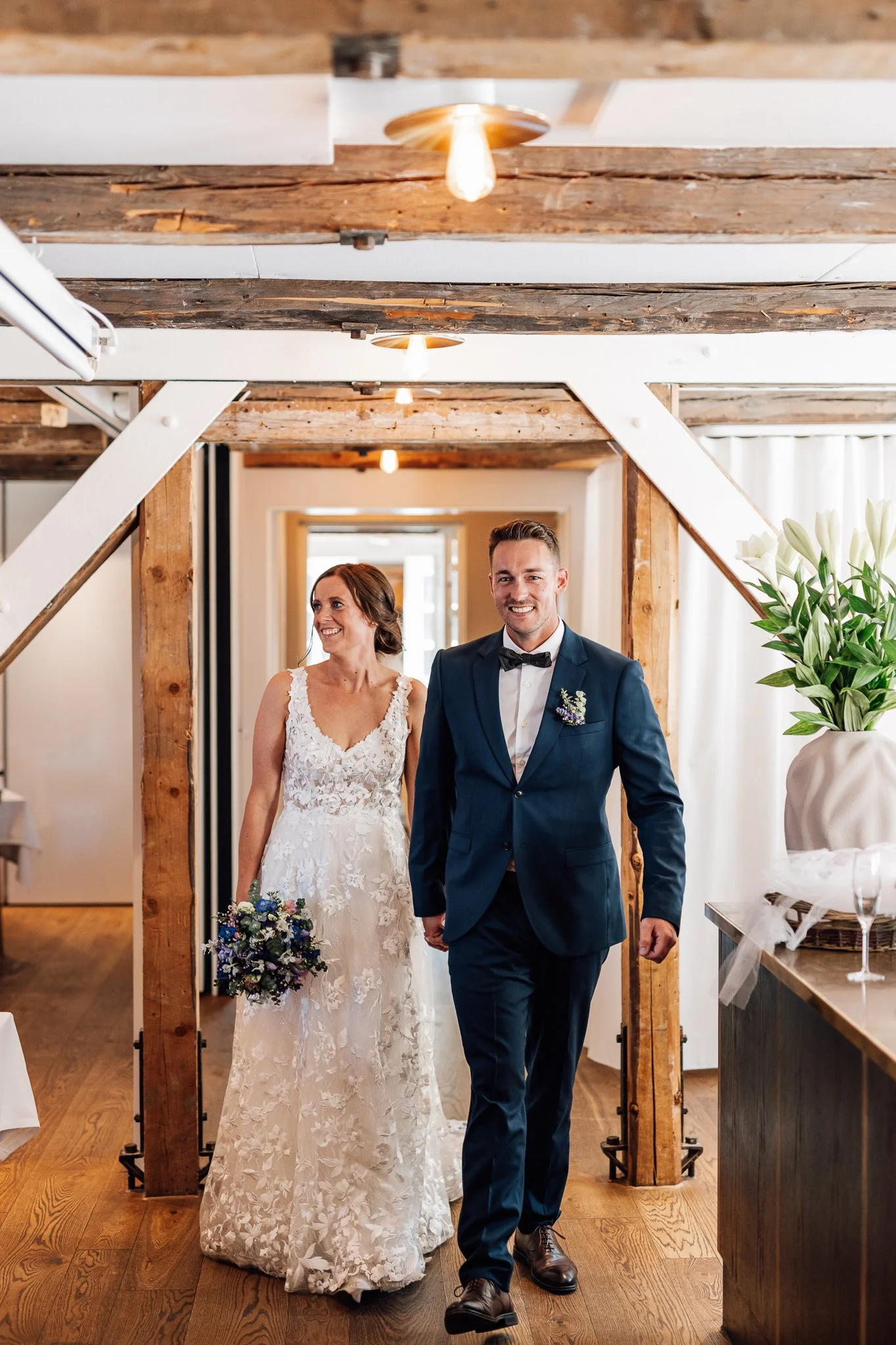 En brud og en brudgom går ind i et bryllupsarrangement, smilende og klædt i formelle gommertøj, i et rum med træbjælker og blomsterdekorationer.