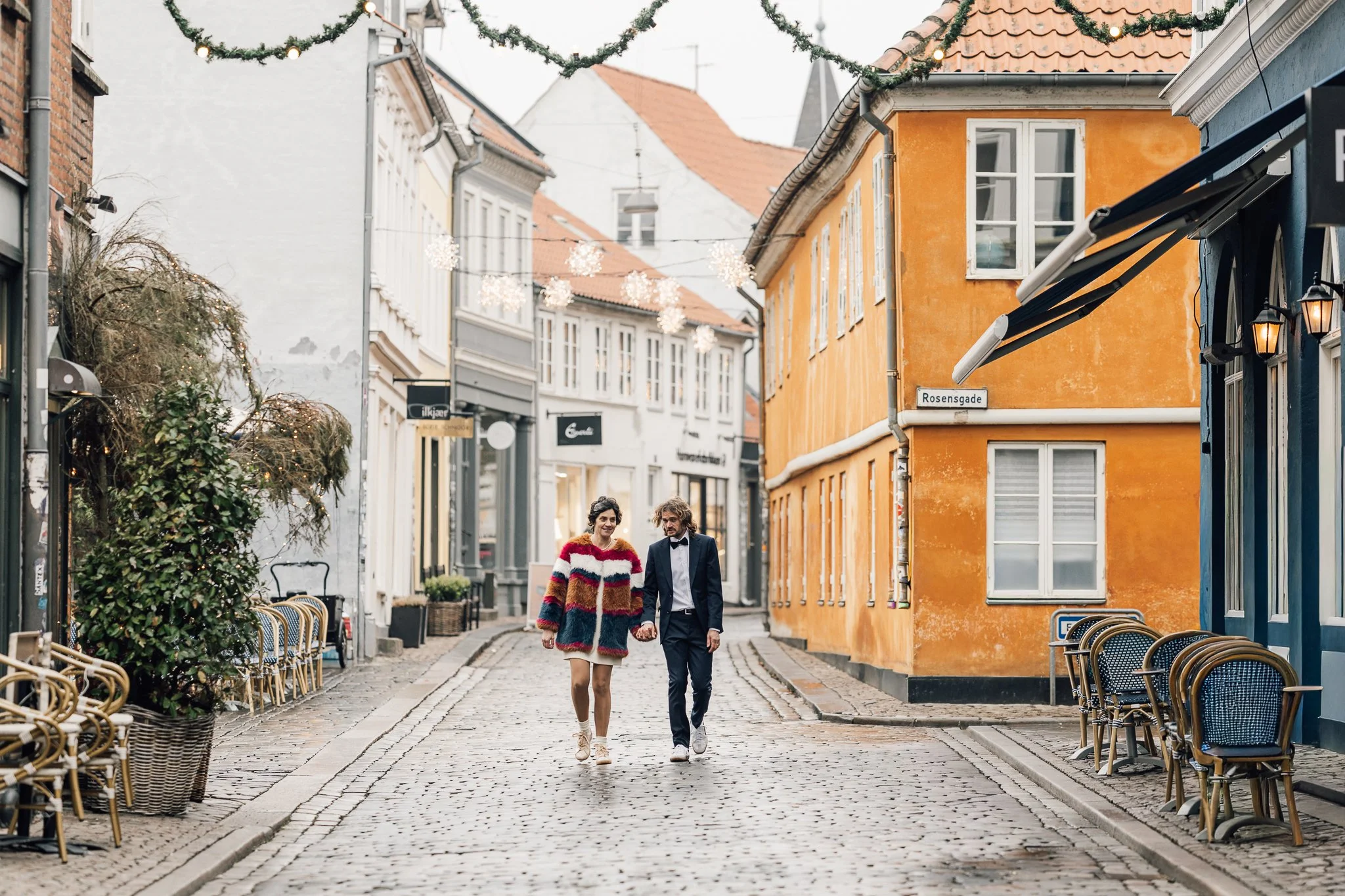 Brudepar går hånd i hånd på en bro i en hyggelig, farverig by med julelys og dekorationer.