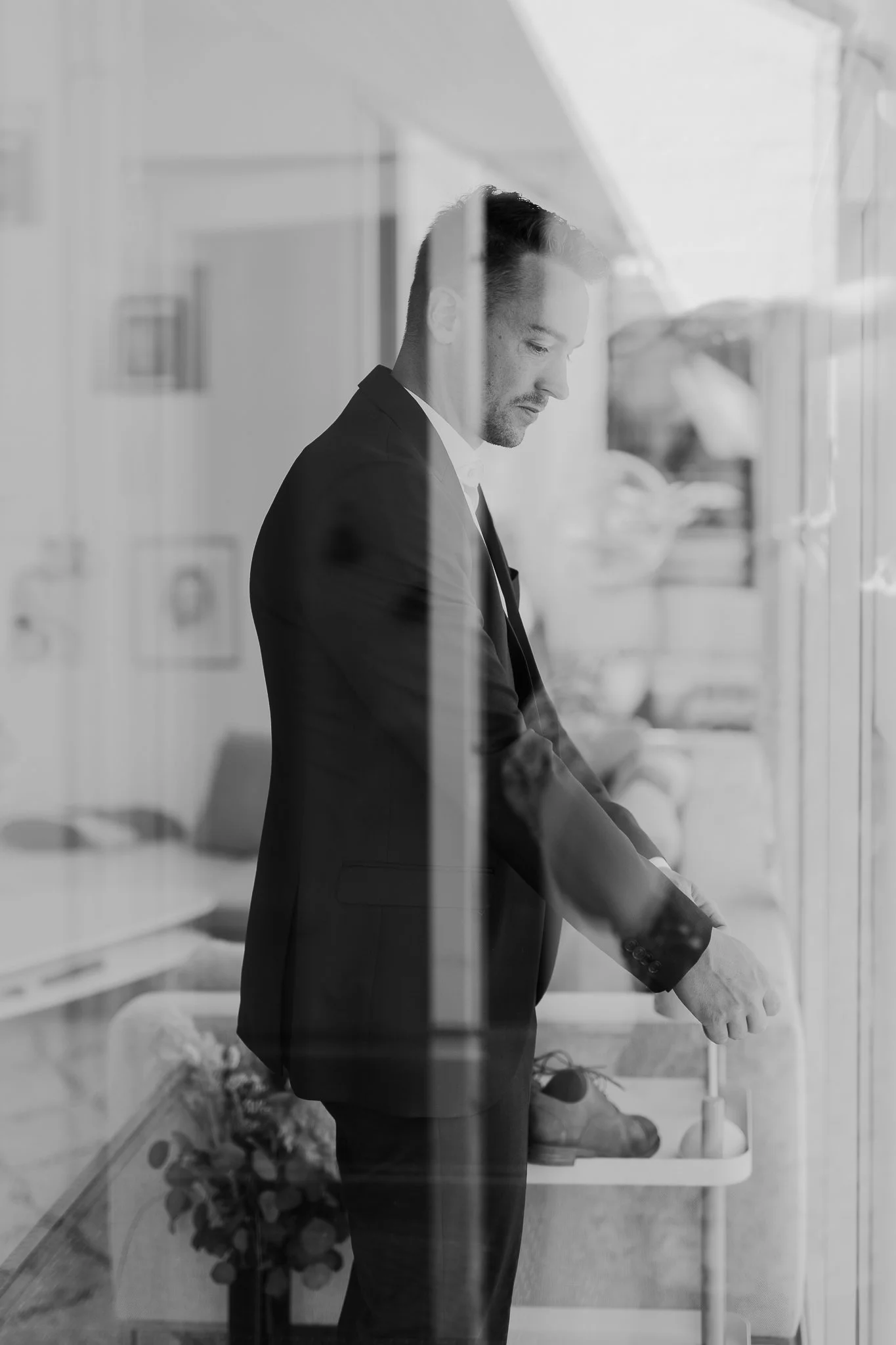 Sort-hvid foto af en mand i mørk habit, der står ved et hospitalssenge med et glas i hånden, gennem en glasrude.