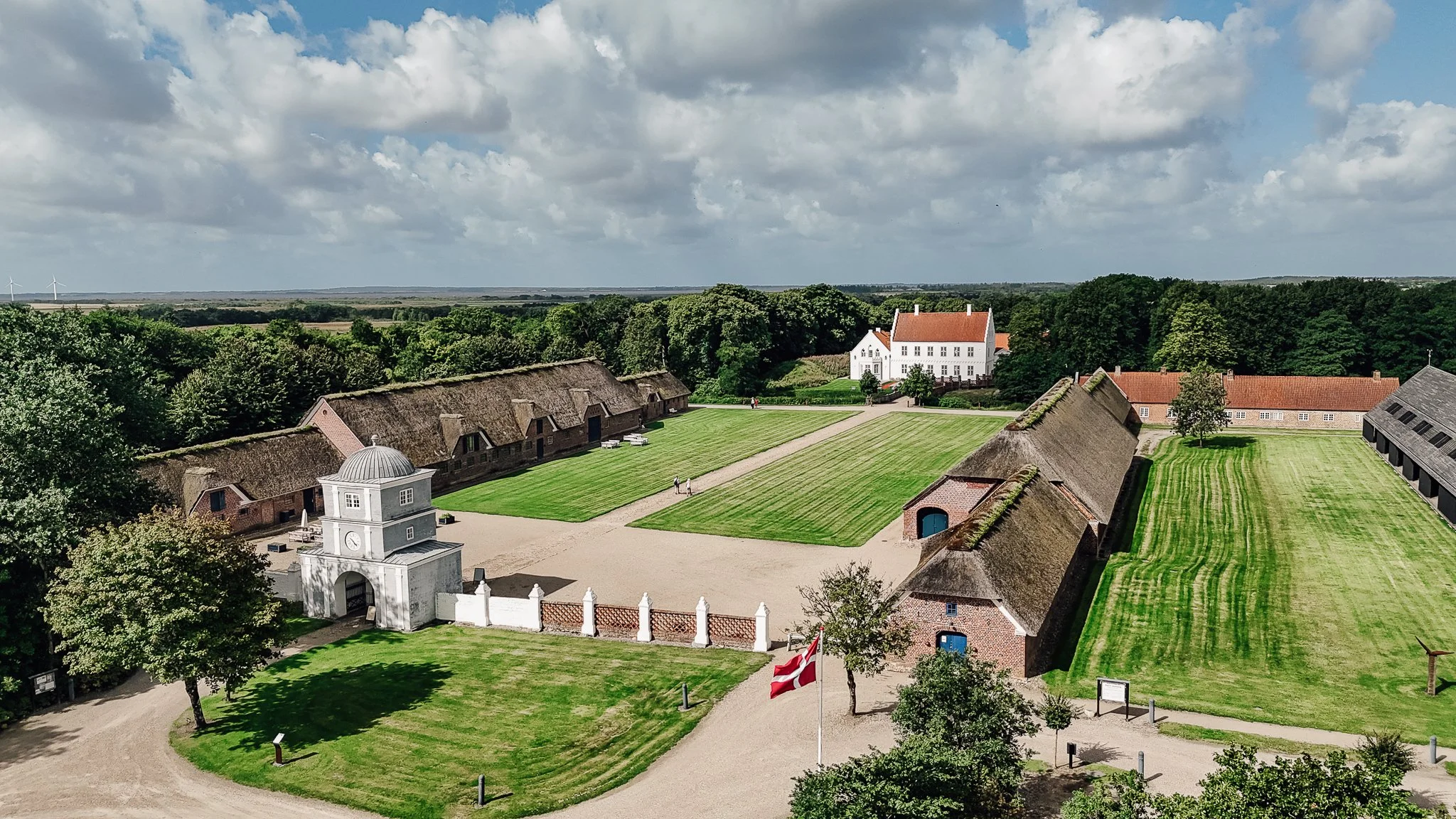 Landskab med græsplæner, historiske bygninger med stråtag, en hvid bygning med klokketårn, nogle træer og en skov i baggrunden under en overskyet himmel.