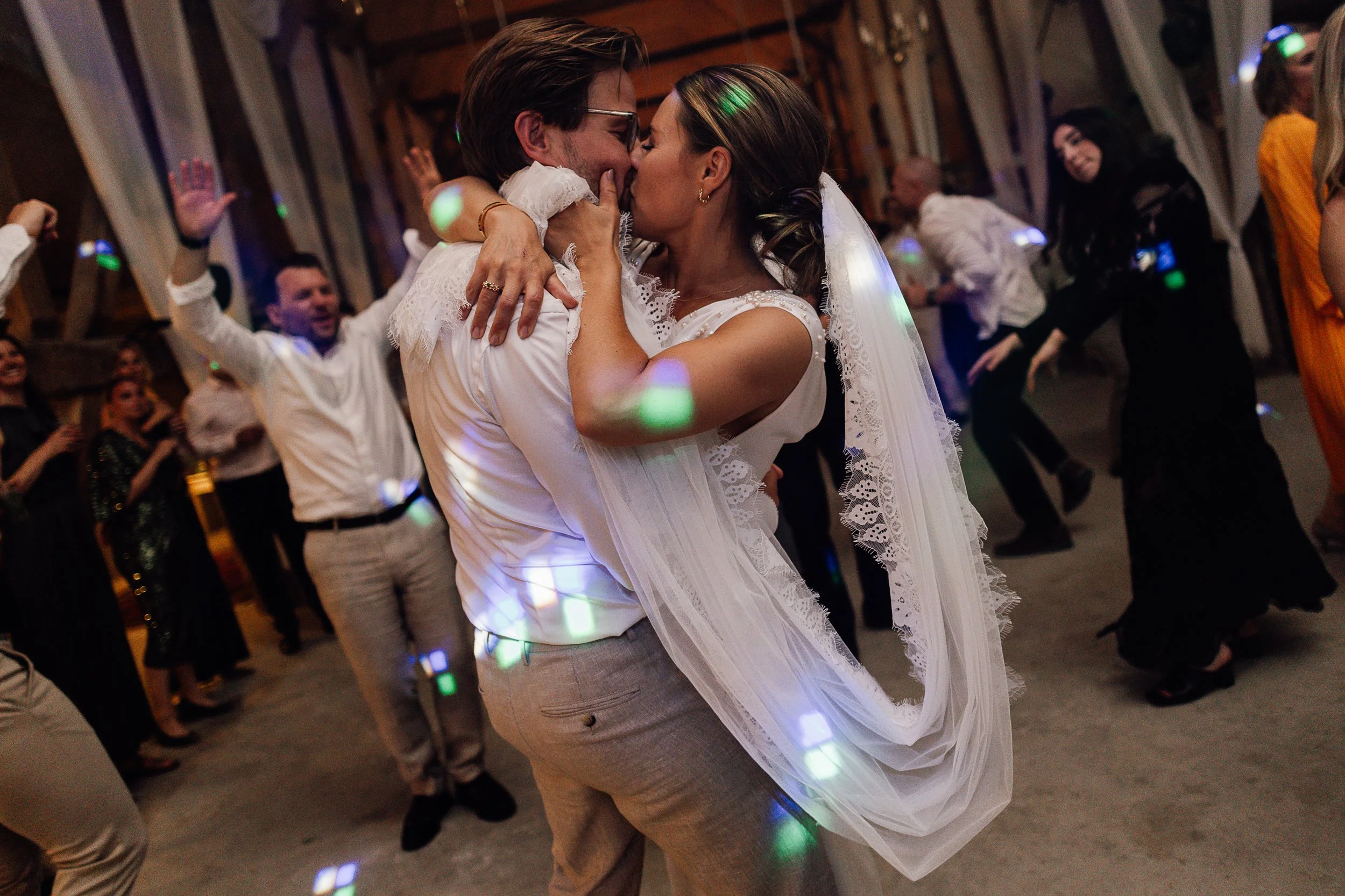 Et brudepar danser tæt sammen, omkranset af mange glade mennesker, i et rustikt trælokale.