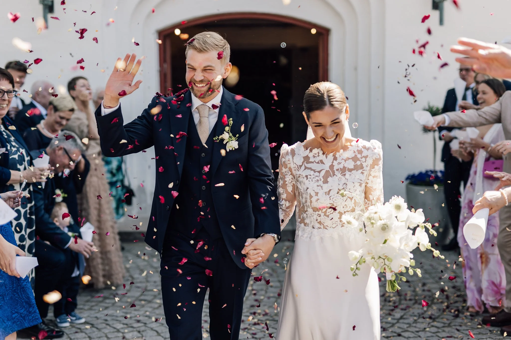 En glad brud og brudgom går hånd i hånd ud af en kirke, mens gæster kaster blomster og konfetti omkring dem.