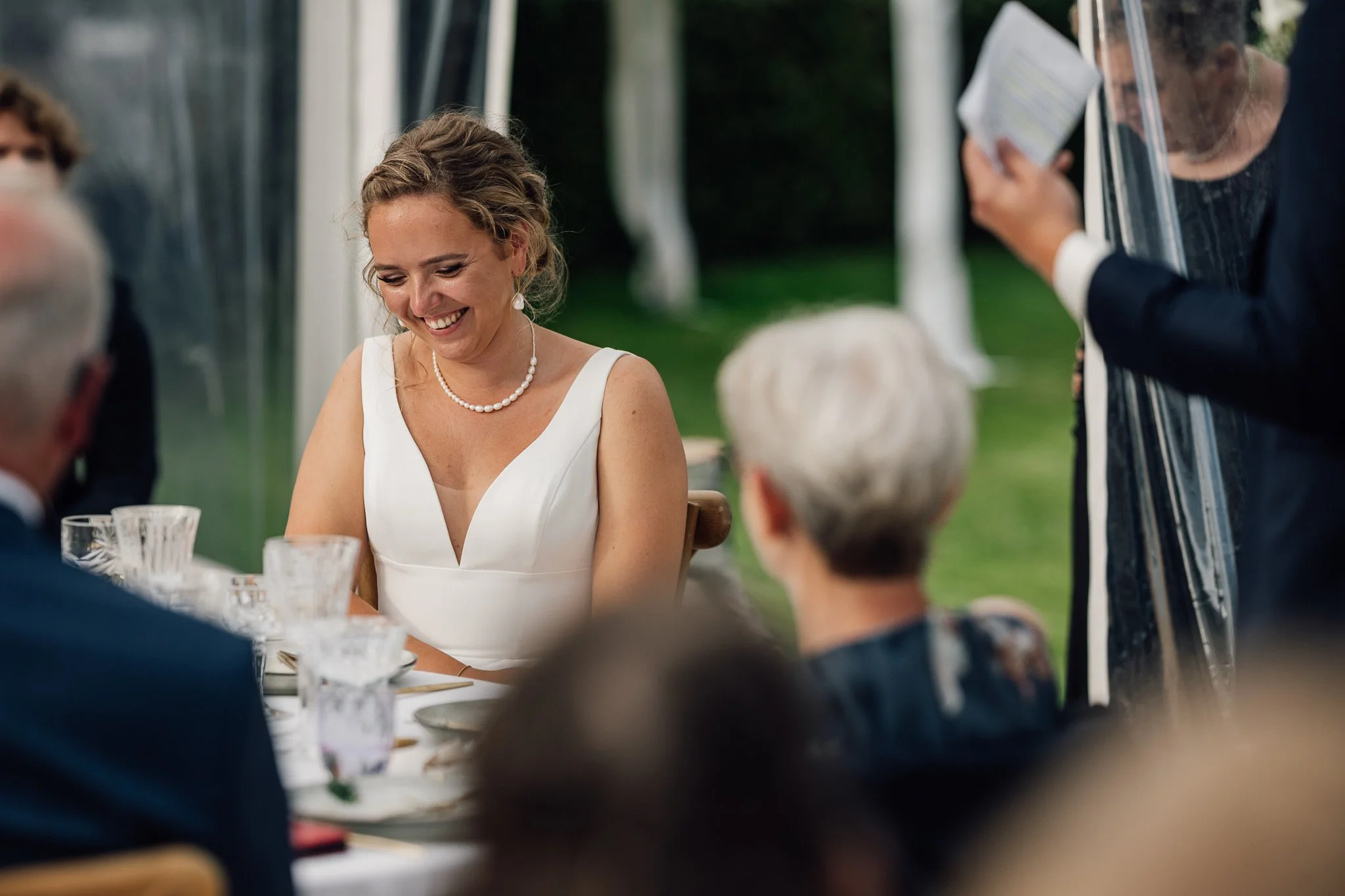 Brud smiler under en udendørs begivenhed med flere mennesker omkring hende.