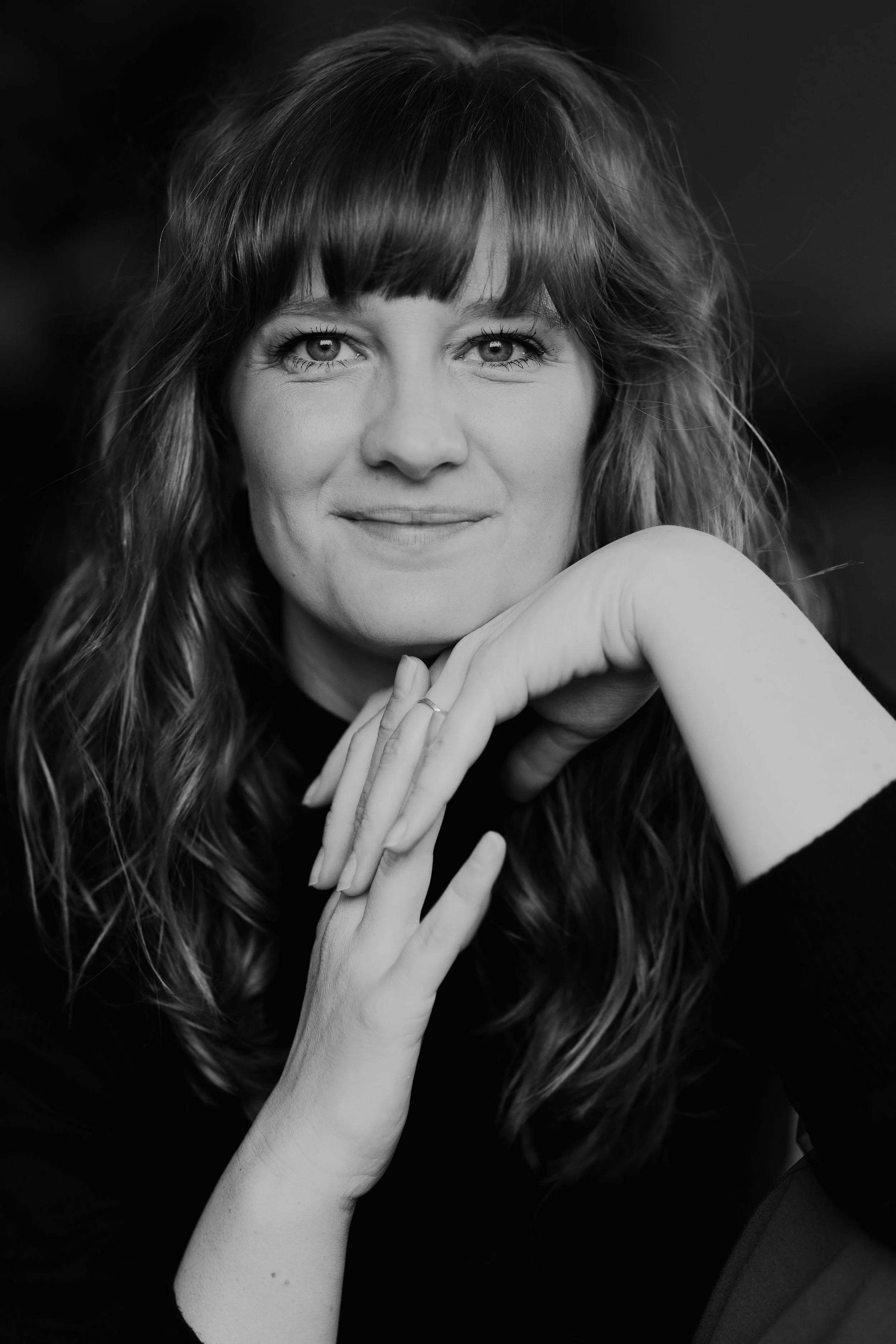 Sort-hvid portrætfotografi af en kvinde med langt, bølget hår og smil, der hviler sit ansigt på sin hånd.