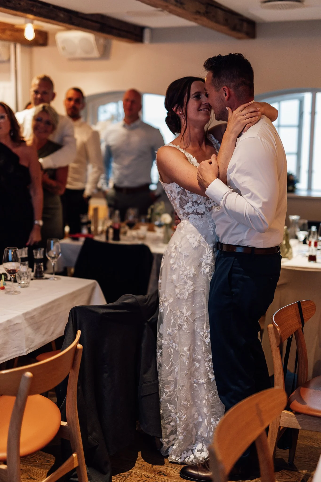 Et brudepar danser tæt sammen i en festlig middagssal, omgivet af gæster, der ser på og smiler.