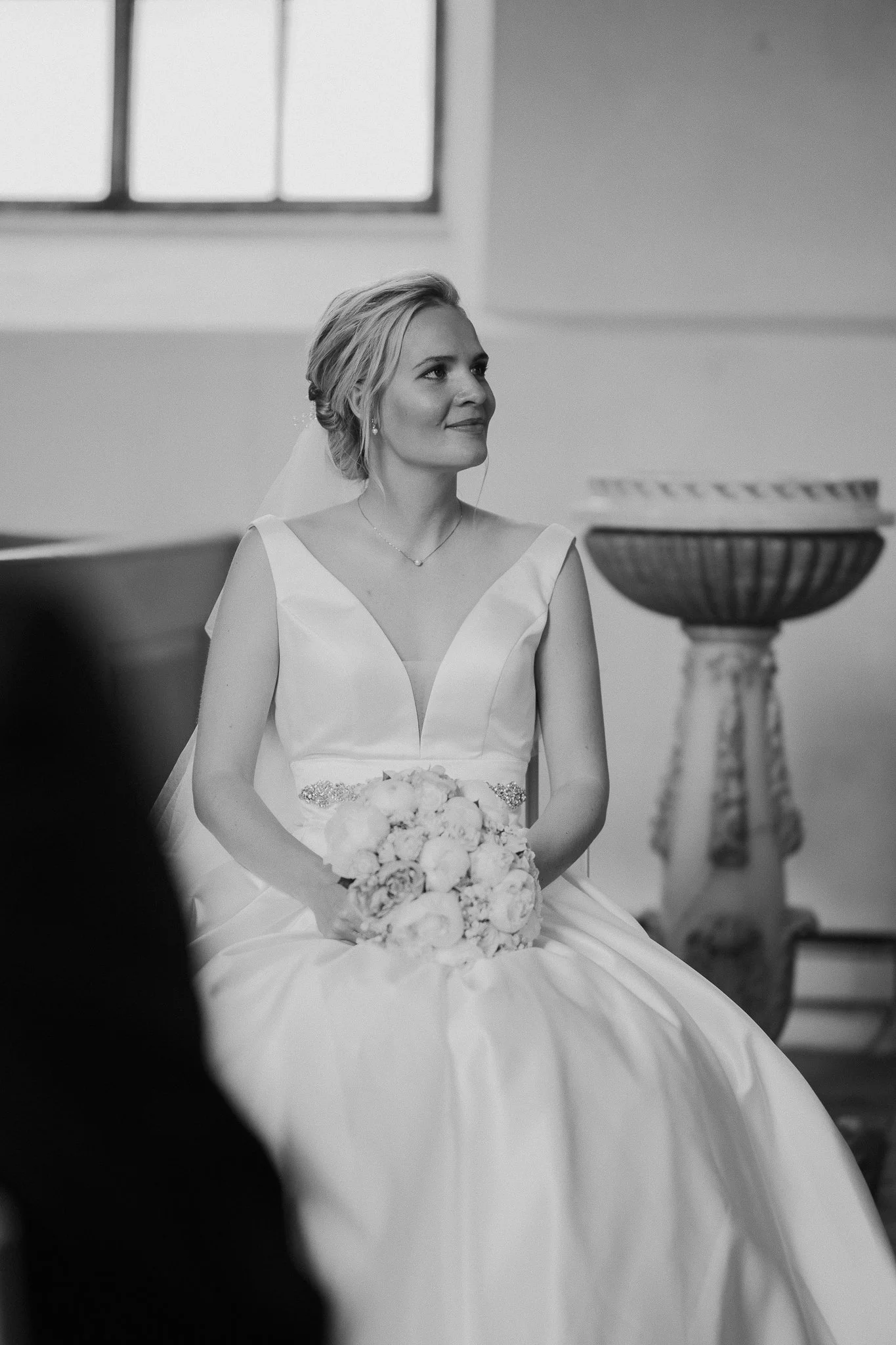 Sort-hvid fotograf af en kvinde i brudekjole, der sidder med en buket blomster i hænderne i en kirke, med et stort vindue i baggrunden og en stor urne på piedestal til højre.
