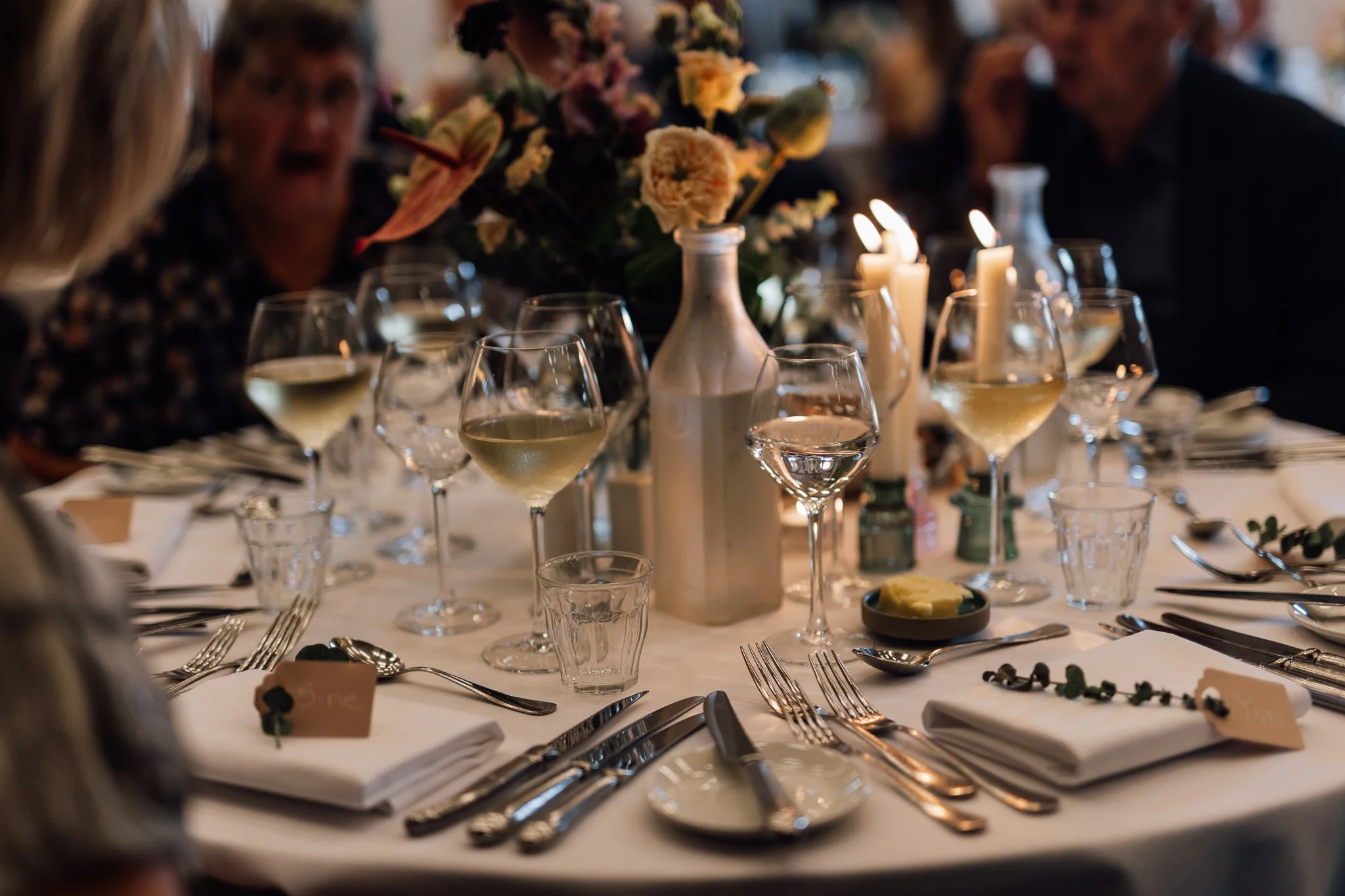 Restaurantbord med glas vin, stearinlys, blomsterdekoration og bestik.