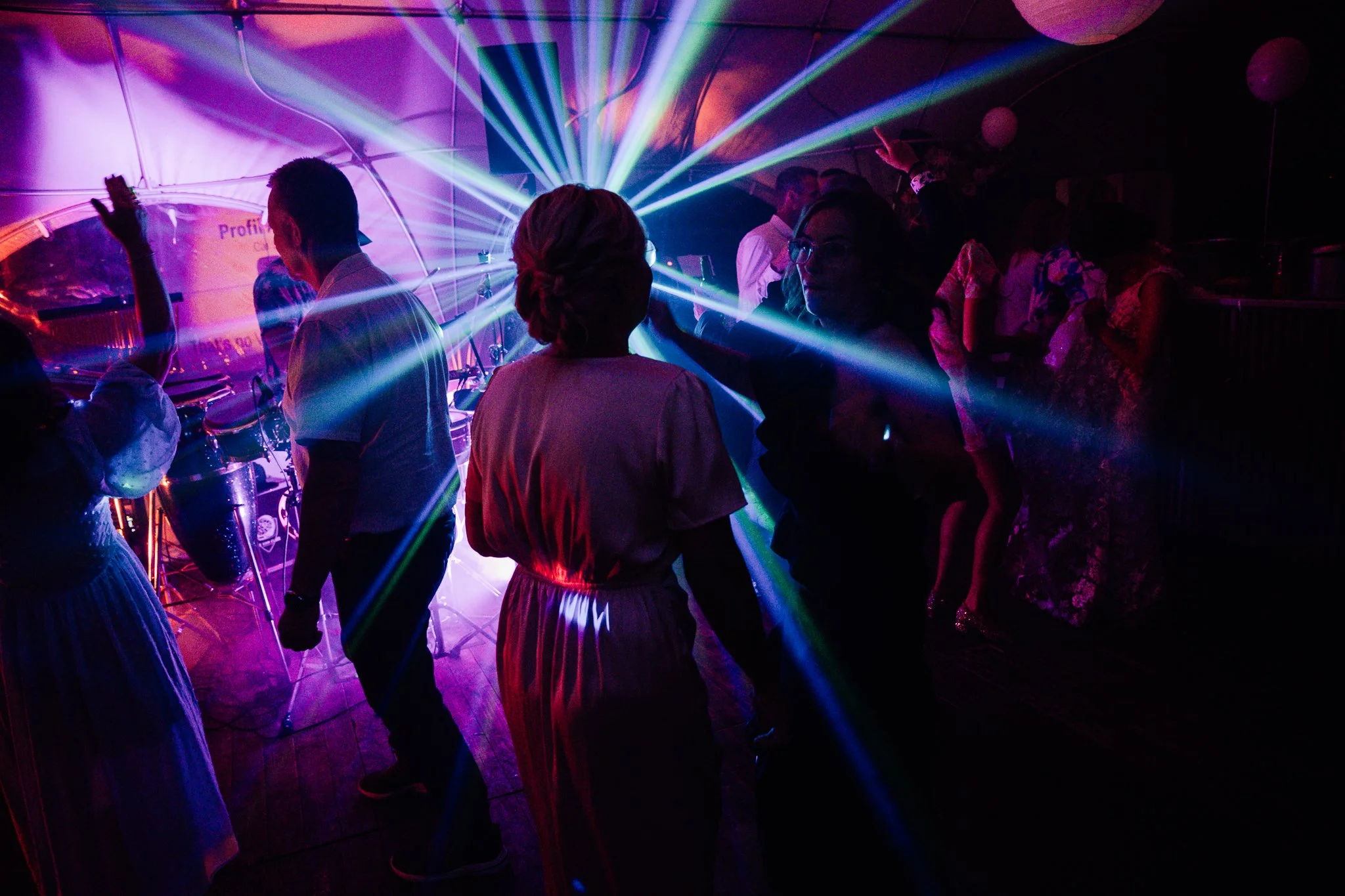 Folk danser og danser til musik under en fest, med farverige lysstråler, dekorationer og balloner i et festligt miljø.