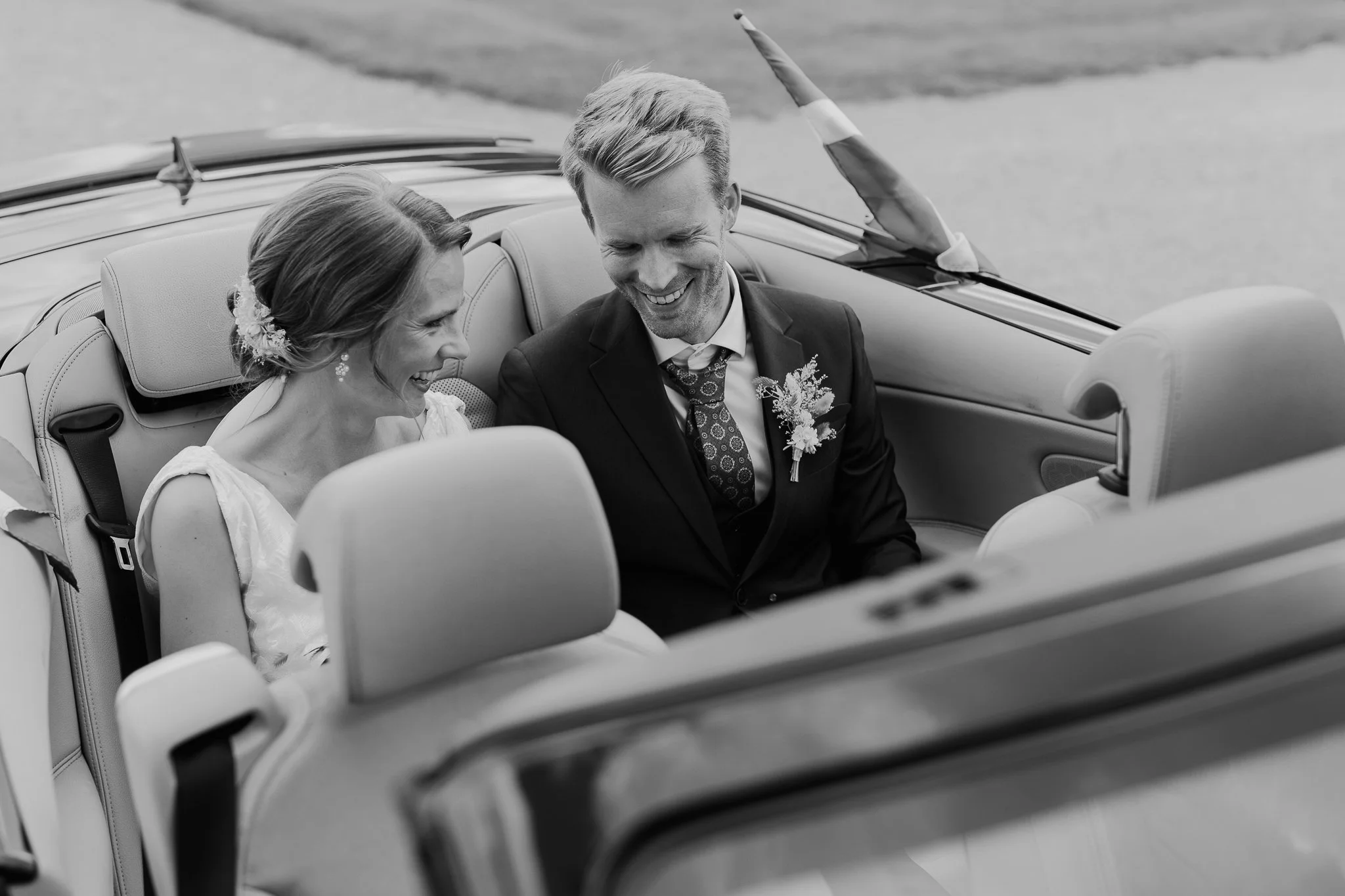 Et brudepar, hvor en brud og en brudgom sidder i en cabriolet, smiler og har en glædelig dag.